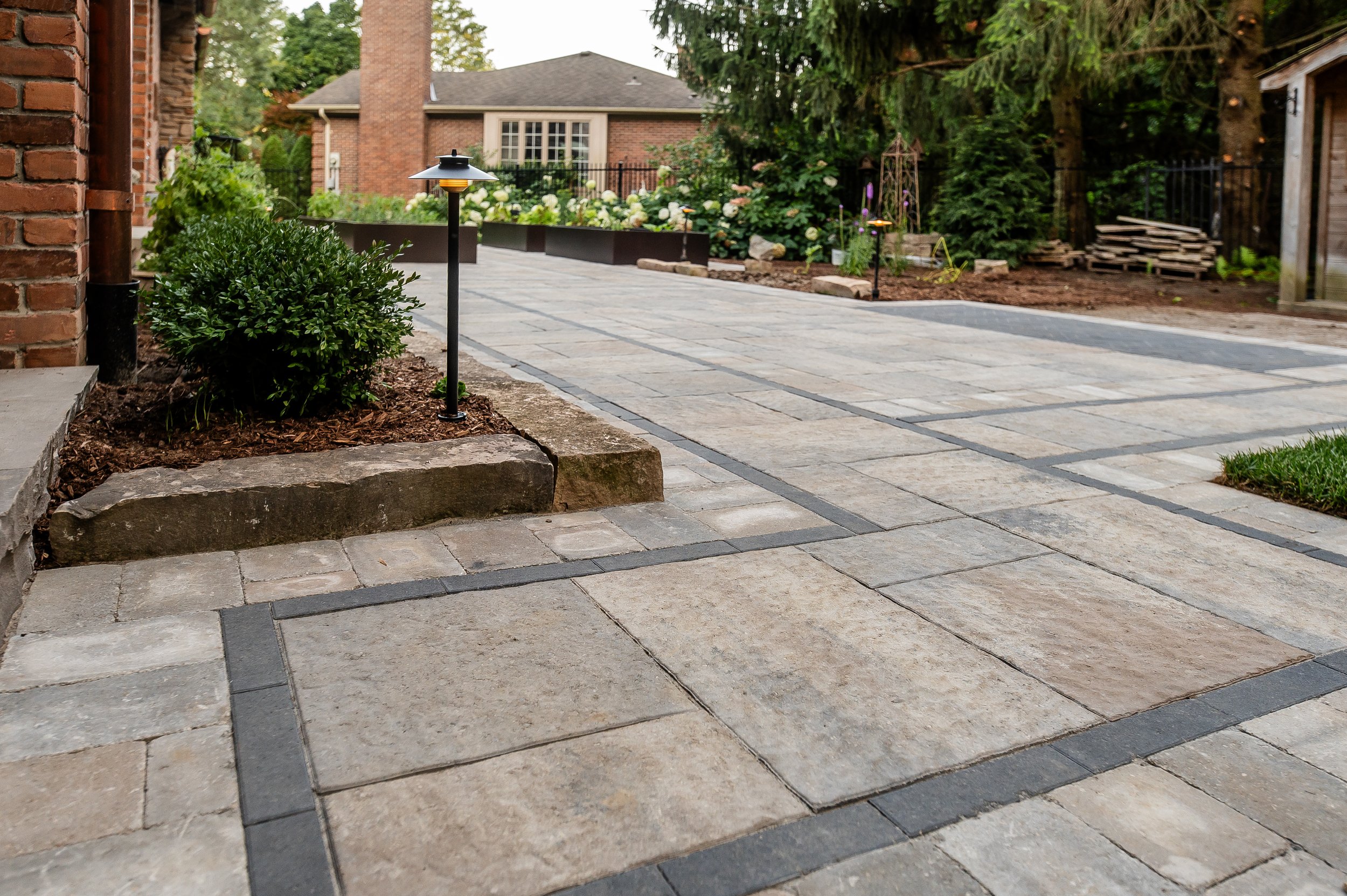 Paved driveway with decorative tiles, bordered by small bushes and garden lights. In the background, there is a brick house with windows and a garden area with trees and plants.