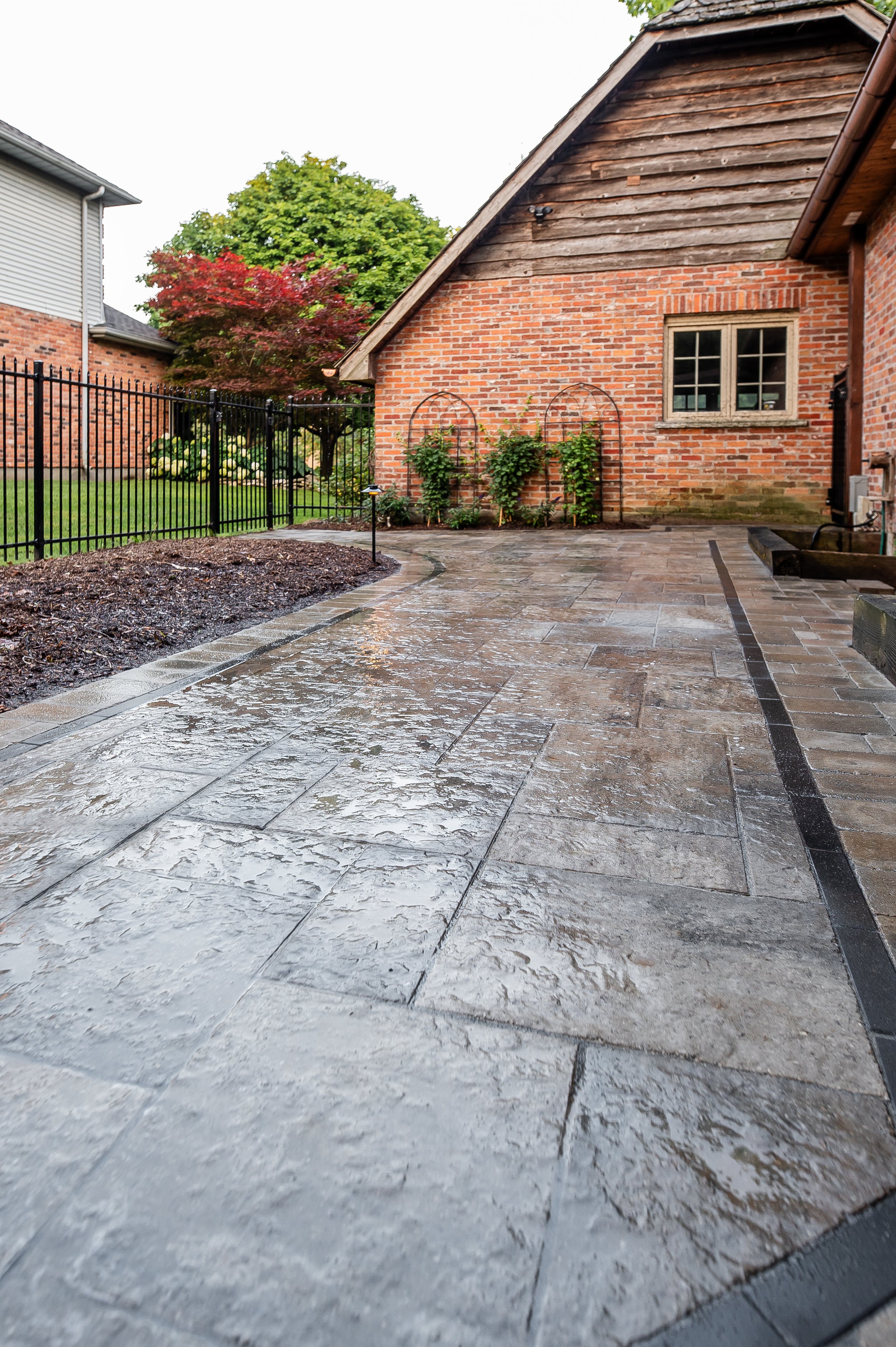 A wet, textured stone patio leads to a brick house with a wooden upper facade and a small window. To the left, there is a black metal fence and garden with green and red foliage. The sky appears overcast, indicating recent rain.