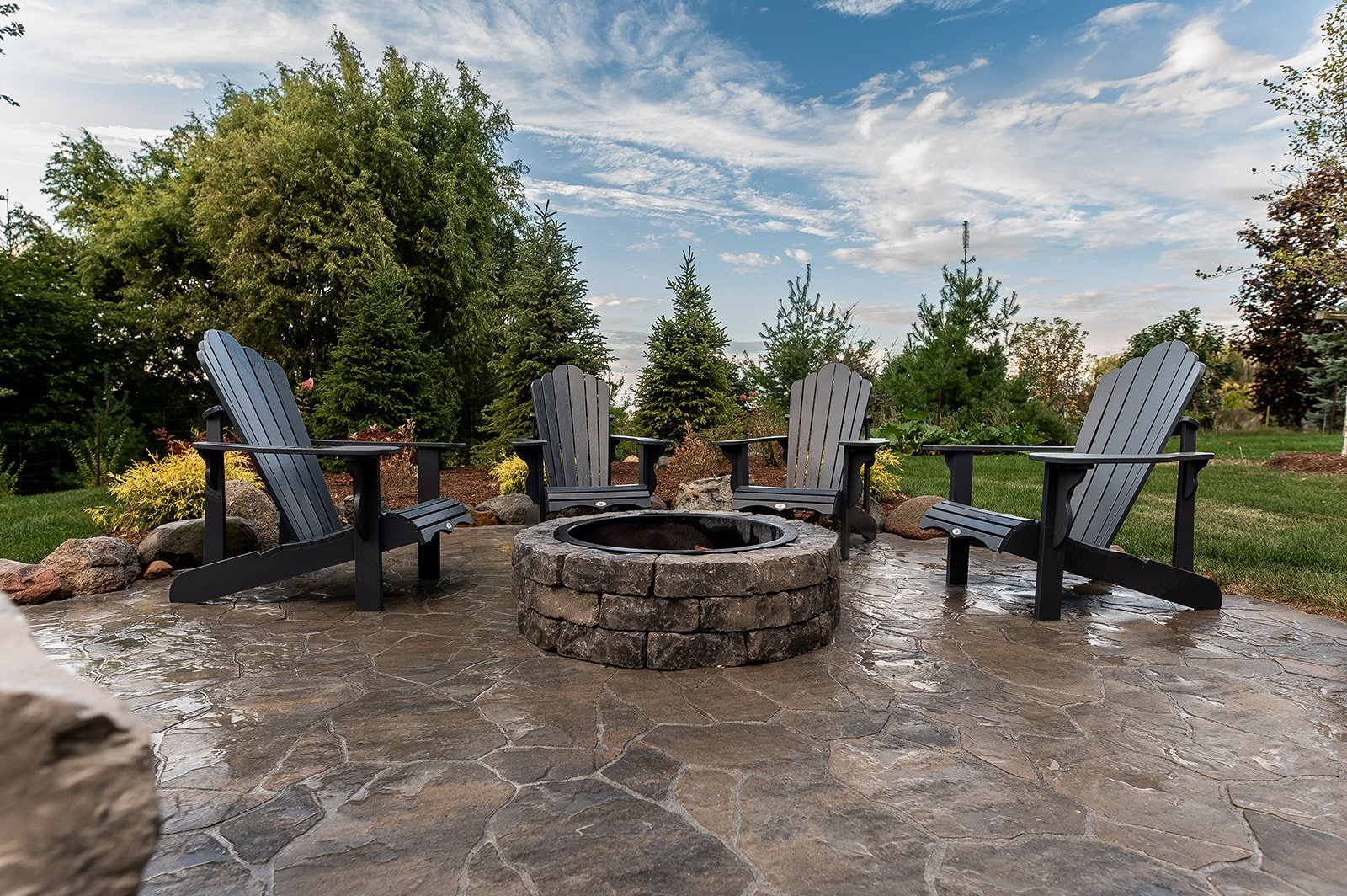Outdoor patio with four Adirondack chairs surrounding a stone fire pit, set on a stamped concrete surface, surrounded by trees and greenery under a partly cloudy sky.
