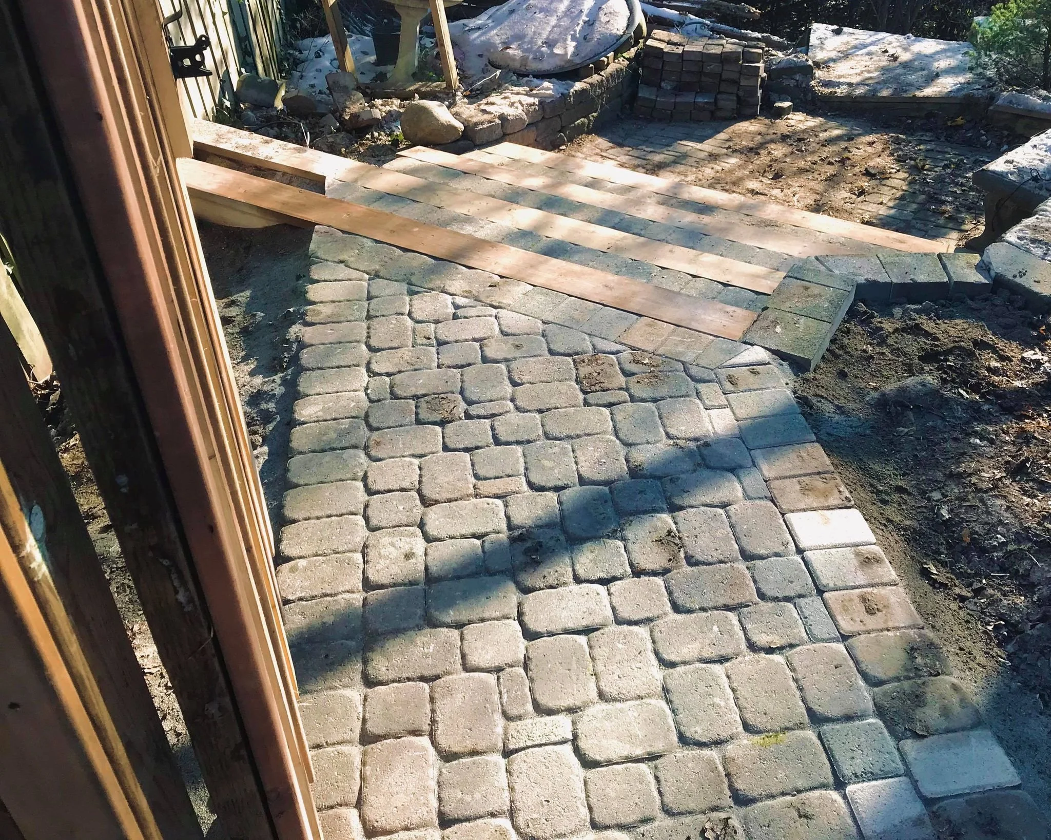 Outdoor stone patio under construction with paving stones, wooden planks, and stacks of bricks.