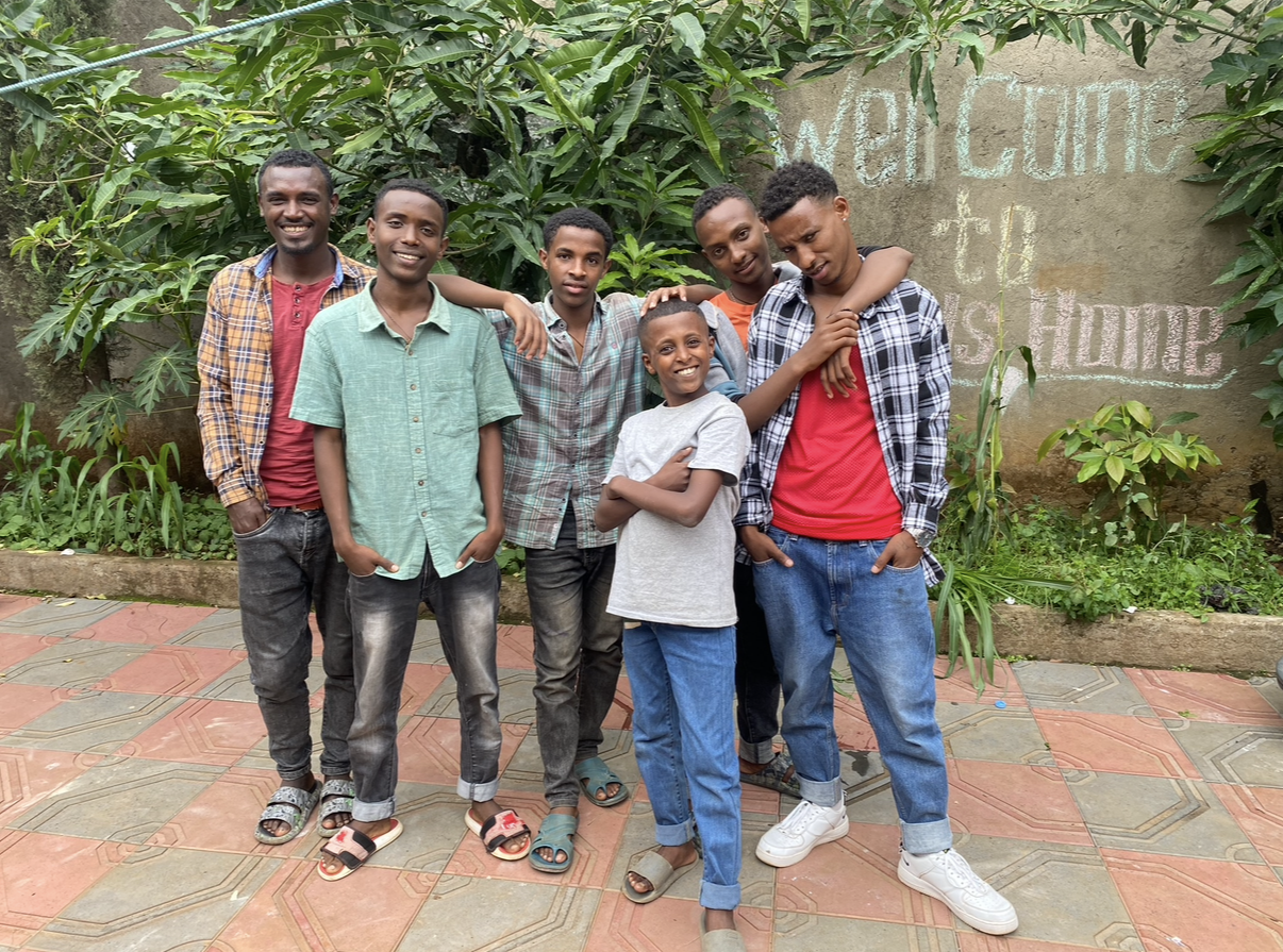 Seven young boys standing together outdoors on a tiled pathway, with a lush green garden and a concrete wall with colorful chalk writing in the background.