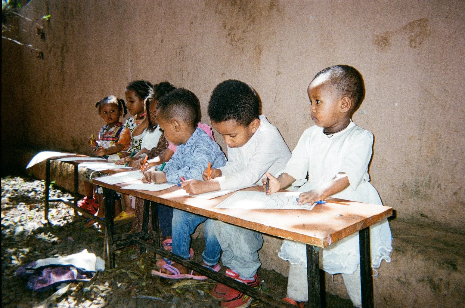 A group of children sitting at a wooden table inside a room, drawing with colored pencils on paper.