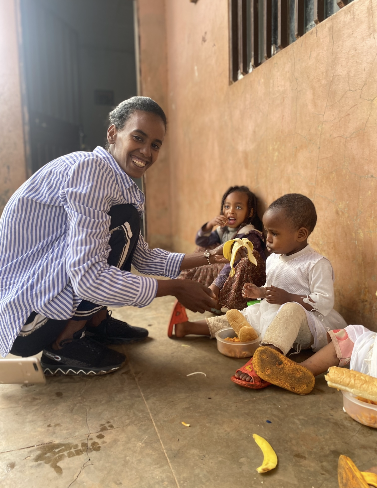 A woman kneeling and smiling offers a banana to two children sitting on the floor against a rust-colored wall. The children are eating snacks and have containers of food in front of them, with one child wearing a white dress and sandals, and the other in a patterned dress with a purple jacket. The scene appears to be outdoors under a covered area.