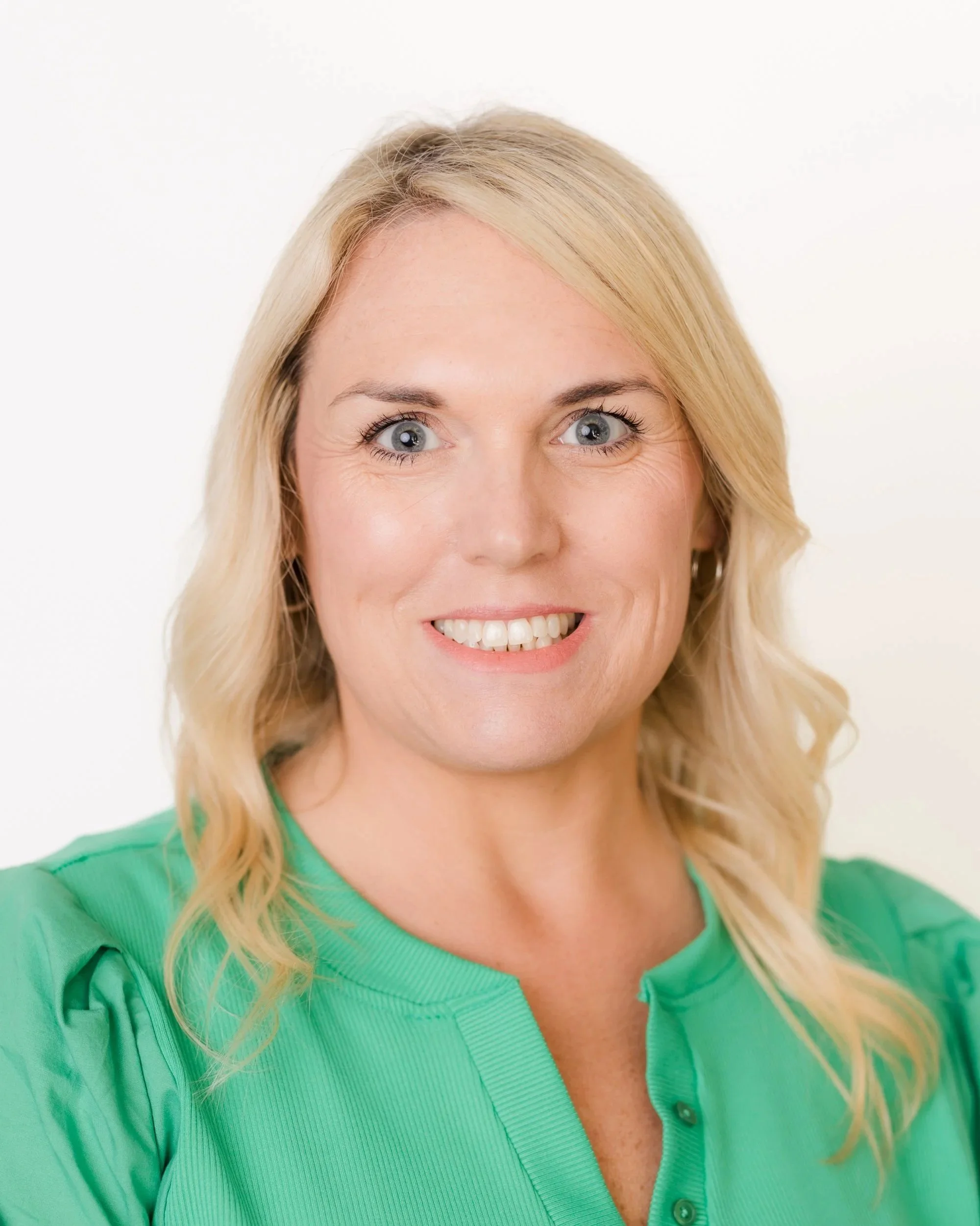 A woman with blonde, wavy hair smiling against a plain gray background.