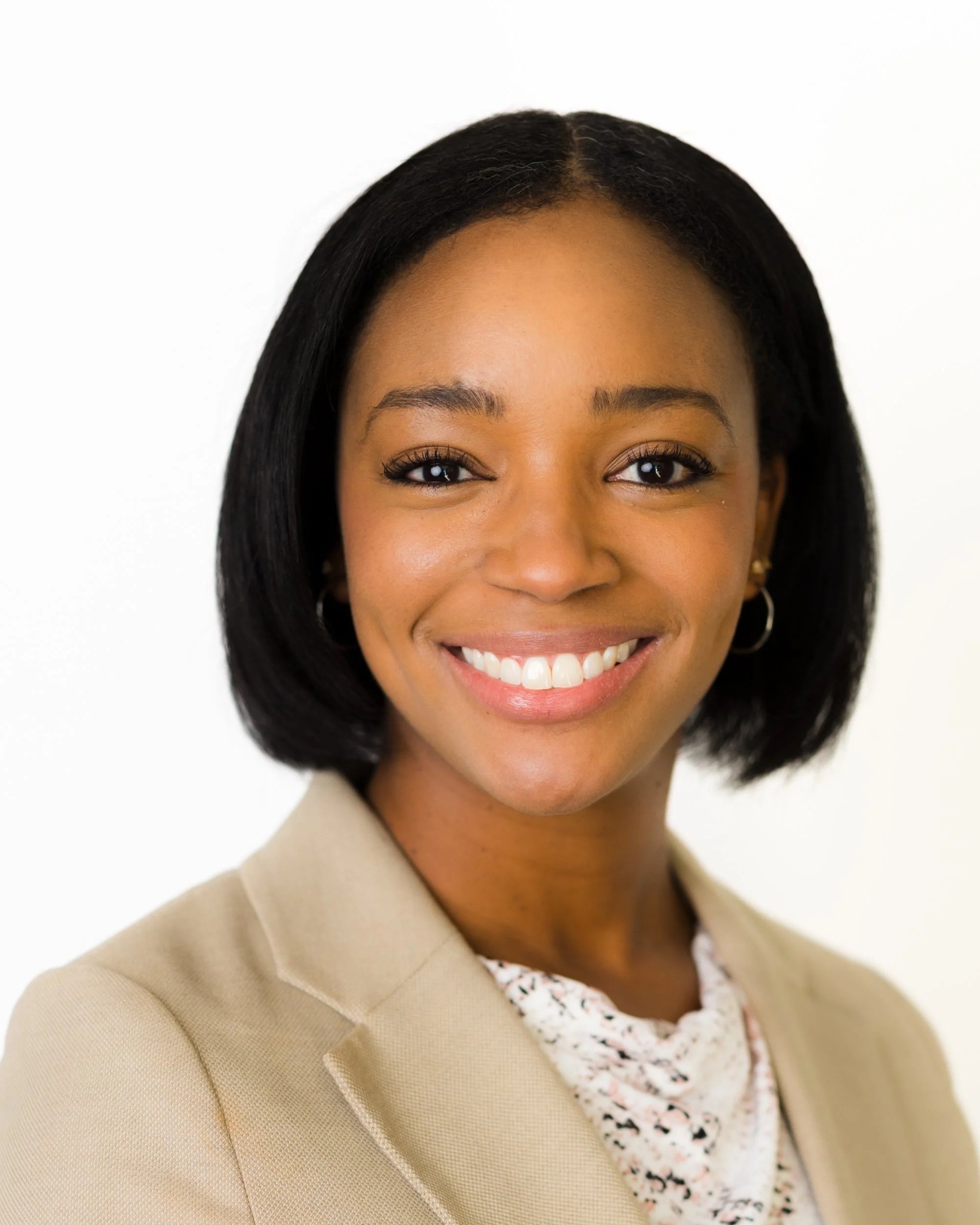 A smiling young woman with straight black hair wearing a white, sleeveless, embroidered top and small hoop earrings against a gray background.