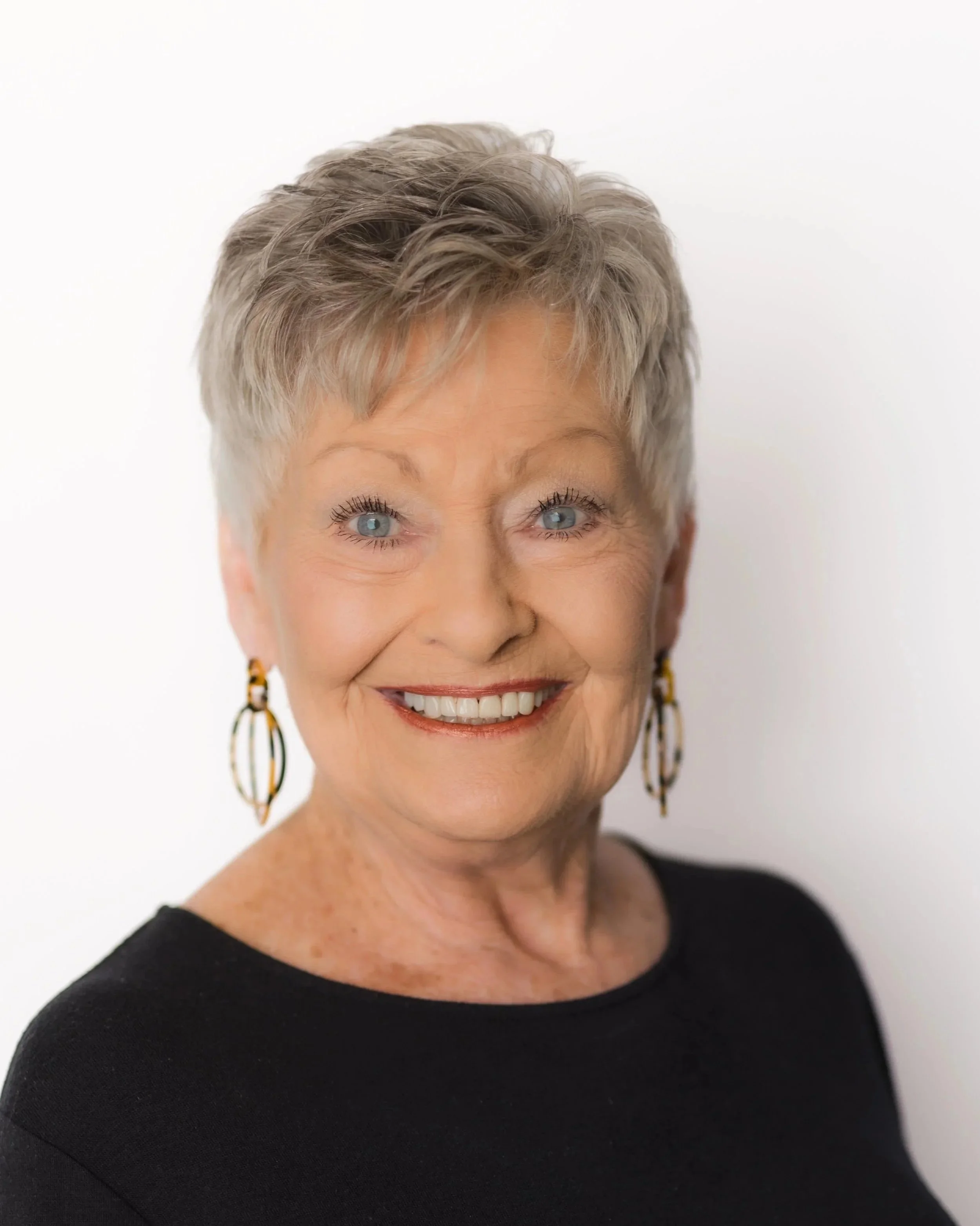 An elderly woman with short gray hair smiling, wearing a black top with ruffled sleeves and earrings, against a plain gray background.