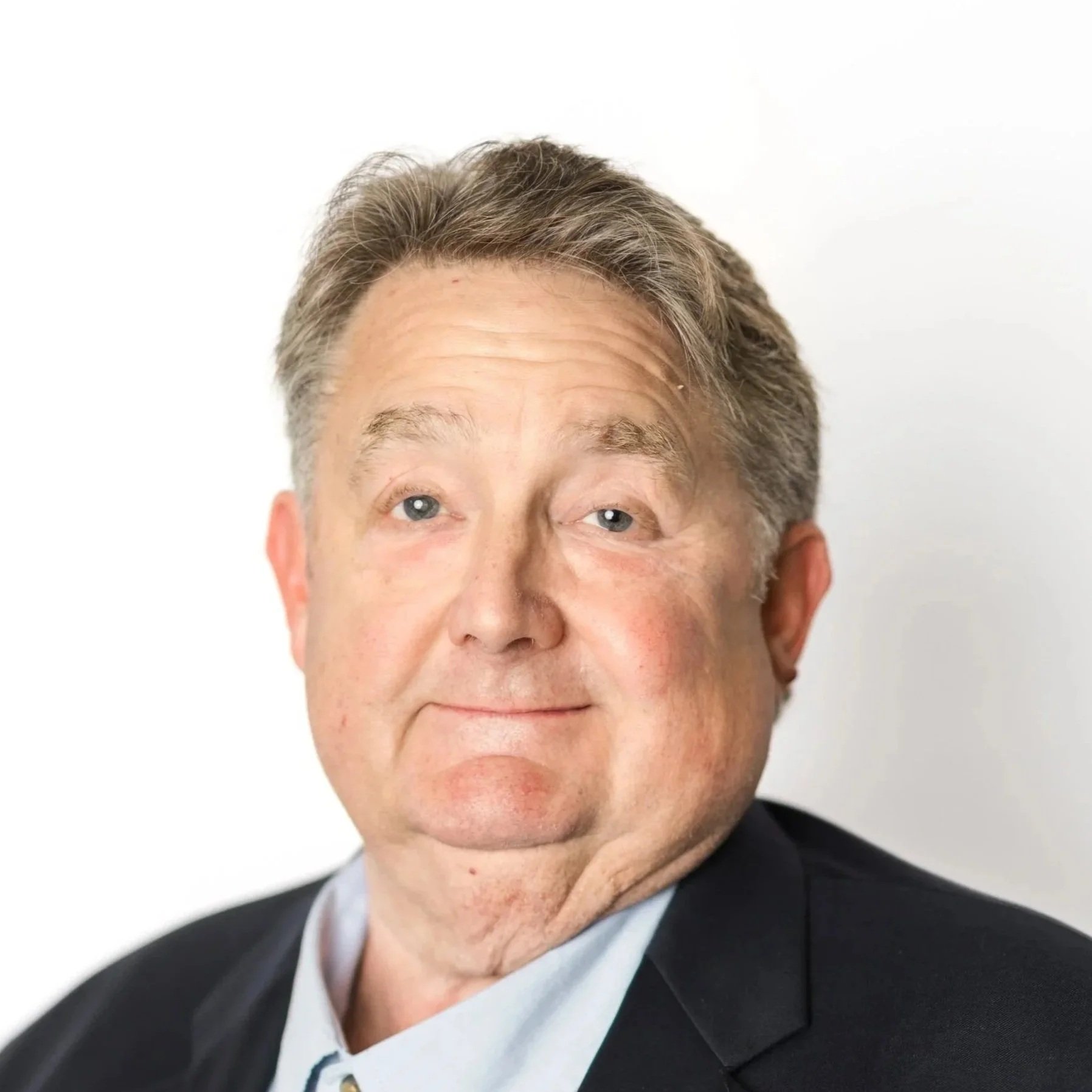 A middle-aged man with brown hair, wearing a black suit, white shirt, and a red, gold, and black patterned tie, sitting against a light gray background.