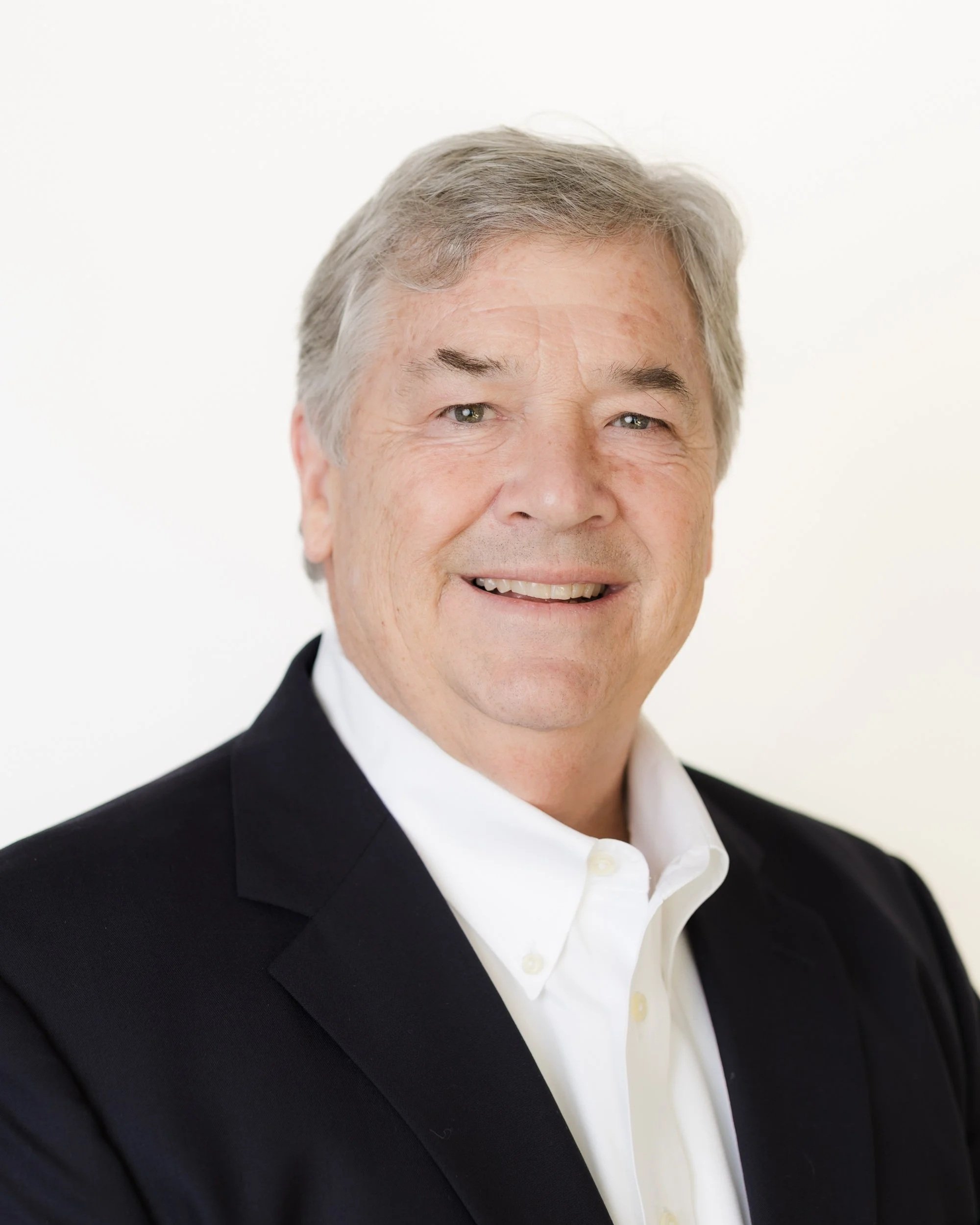 Portrait of a smiling middle-aged man with gray hair, wearing a dark gray jacket and a collared shirt, against a plain gray background.