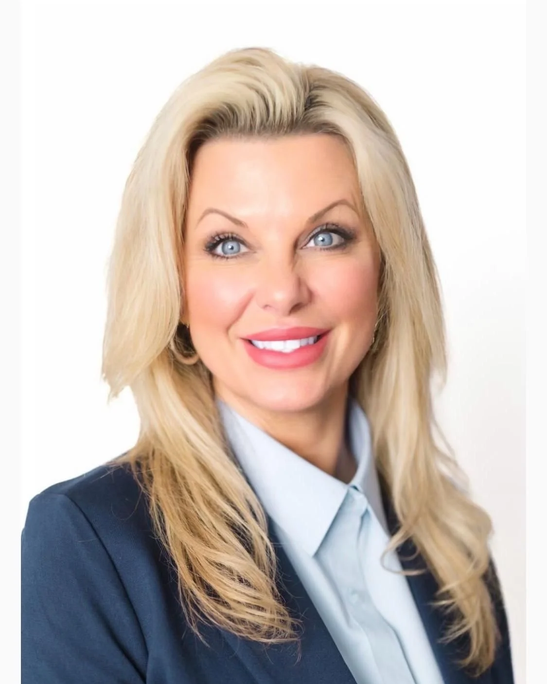 A woman with long, curly blonde hair smiling, wearing a dark blazer over a white top, standing against a plain gray background.