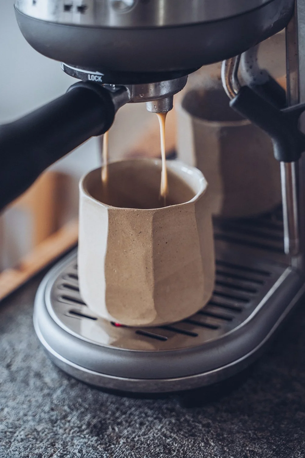 Close-up of espresso being brewed from a coffee machine into a beige, faceted ceramic mug.