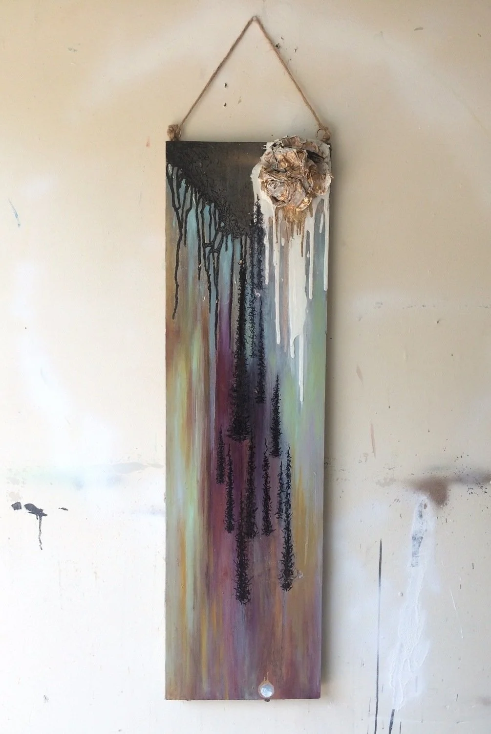 A vertical wall hanging art piece with a dreamcatcher-like decoration made from shells or similar material attached at the top right corner, featuring black, blue, green, purple, and yellow dripping paint effects.