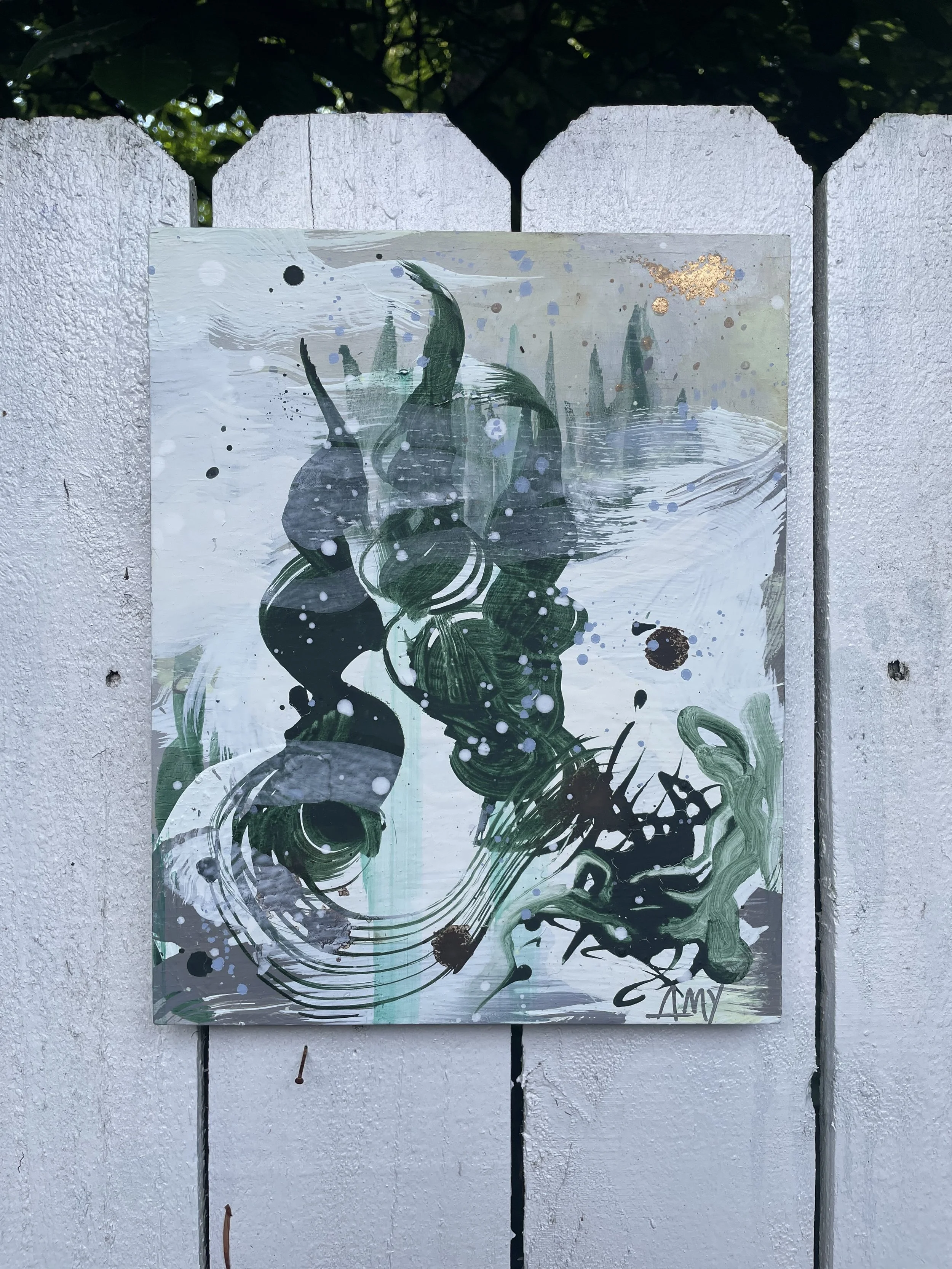 Abstract painting with green, black, white, and gold splashes, brushstrokes, and swirling patterns, signed 'Amy' in the bottom right corner, mounted on a white wooden fence.