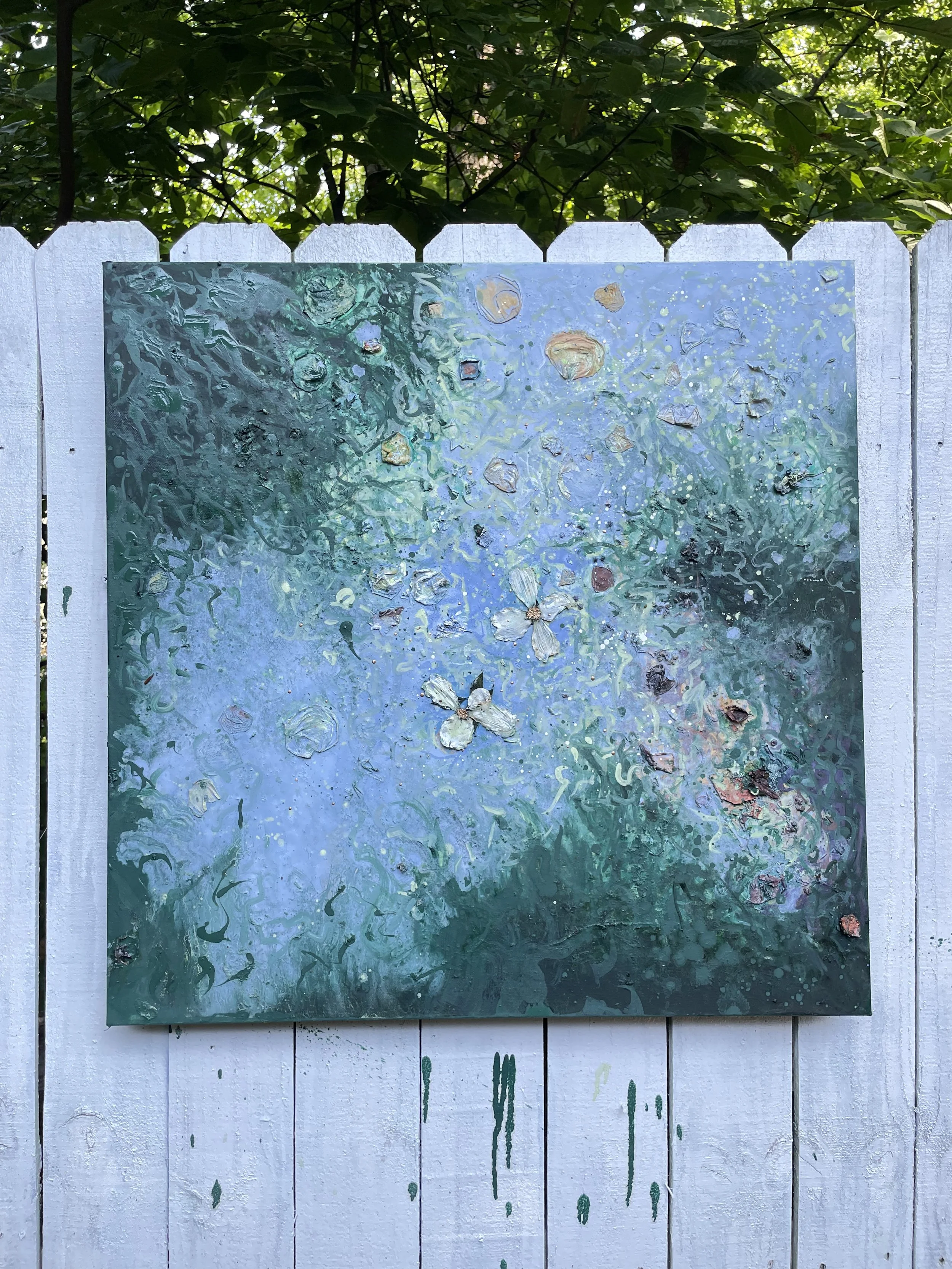 Abstract painting on a white wooden fence with green and blue swirls and floating flower shapes, outdoors with leafy trees in sunlight.