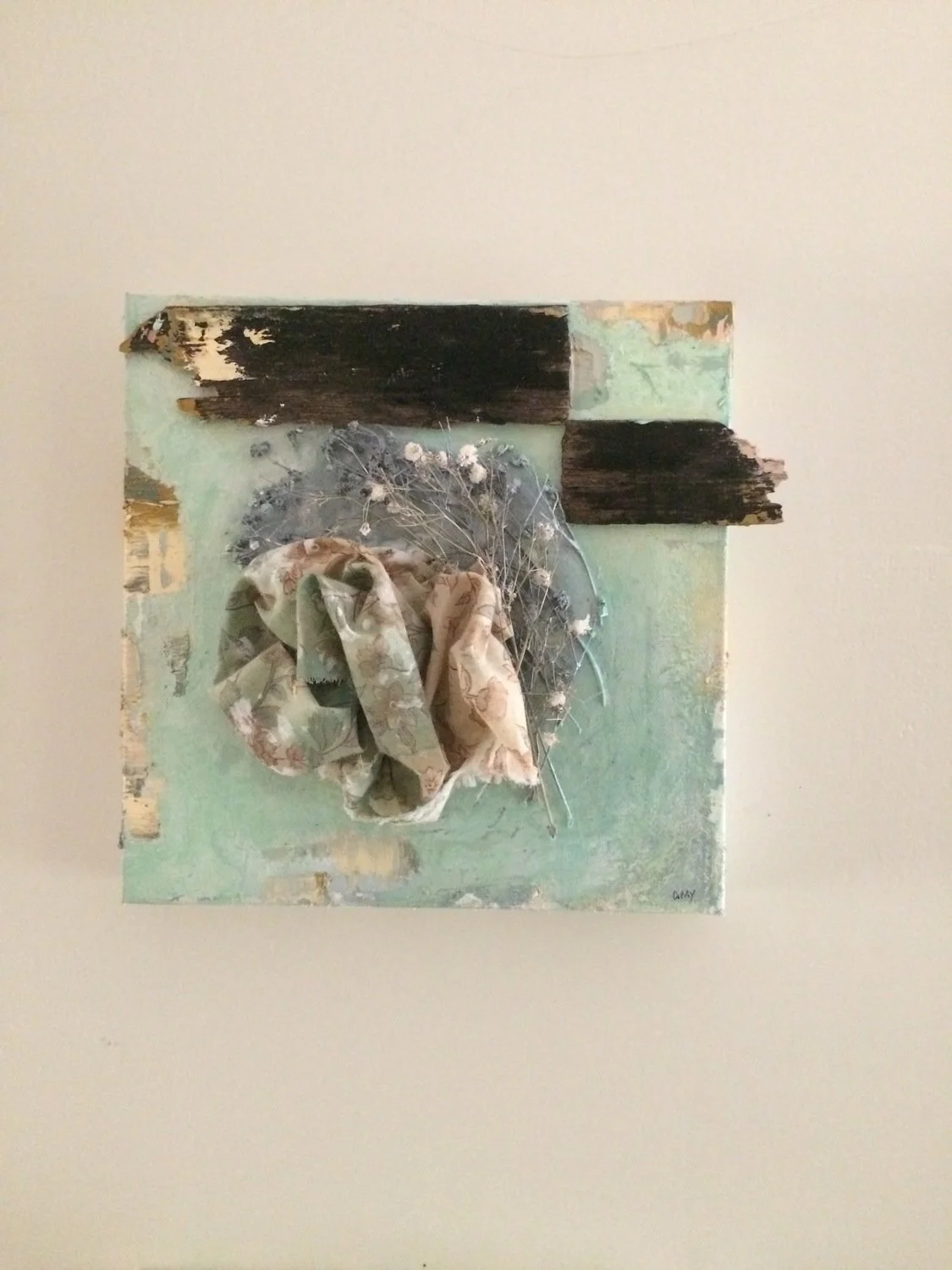 Mixed media artwork featuring distressed wood pieces, a floral-patterned cloth, dried flowers, and layered paint on a square canvas.