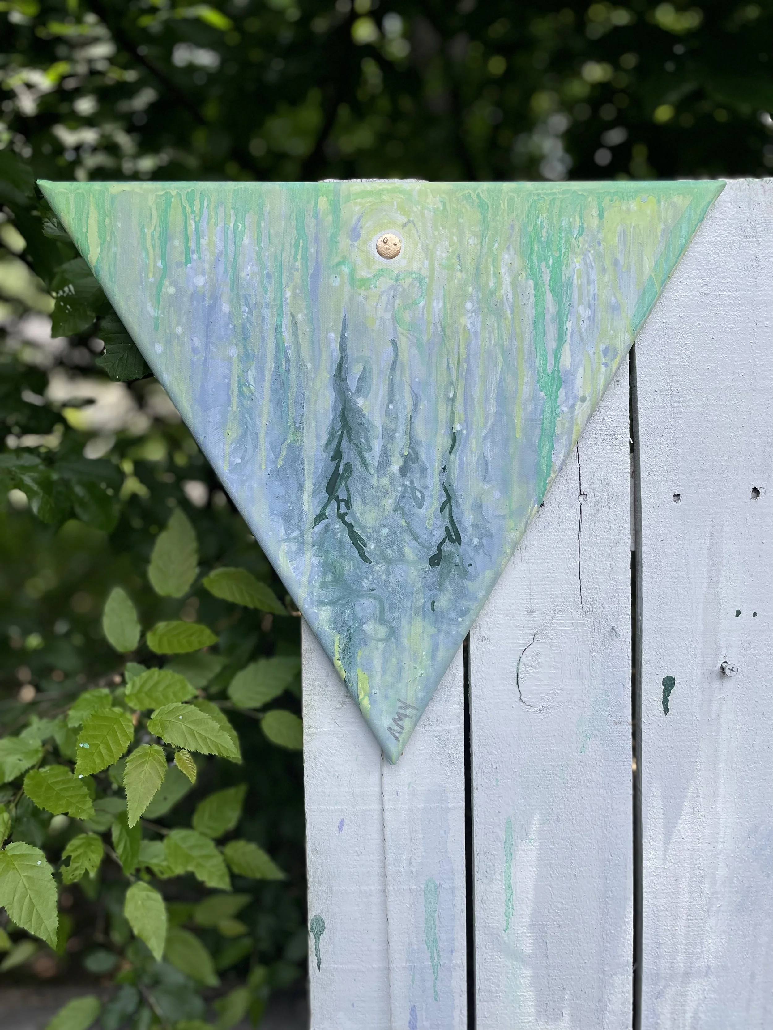 A painted wooden triangle with a moon and tree design, mounted on a white wooden fence with green leaves in the background.