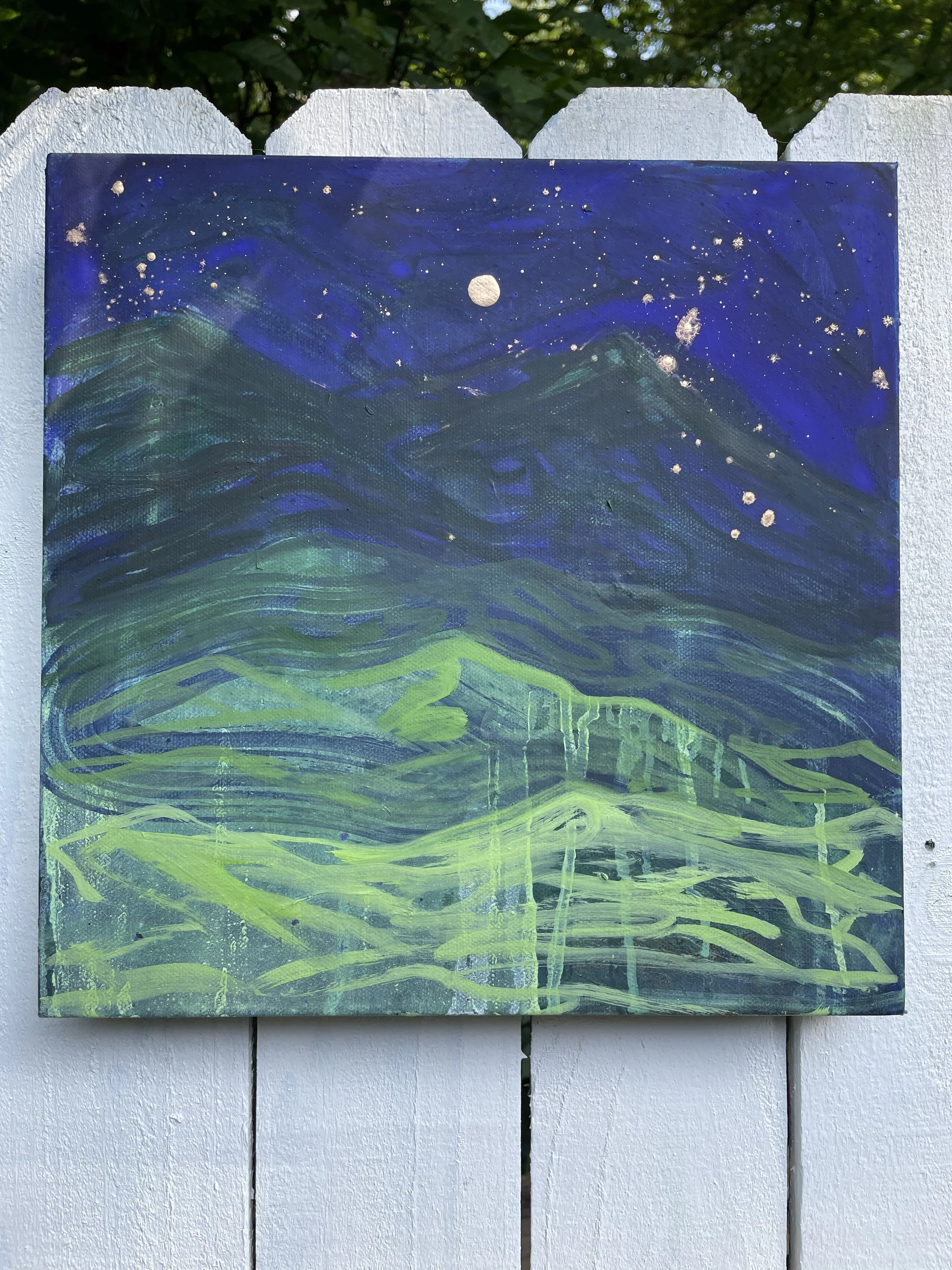 A painting of a night sky with stars, a full moon, and a mountainous landscape illuminated in green, mounted on a white wooden fence.