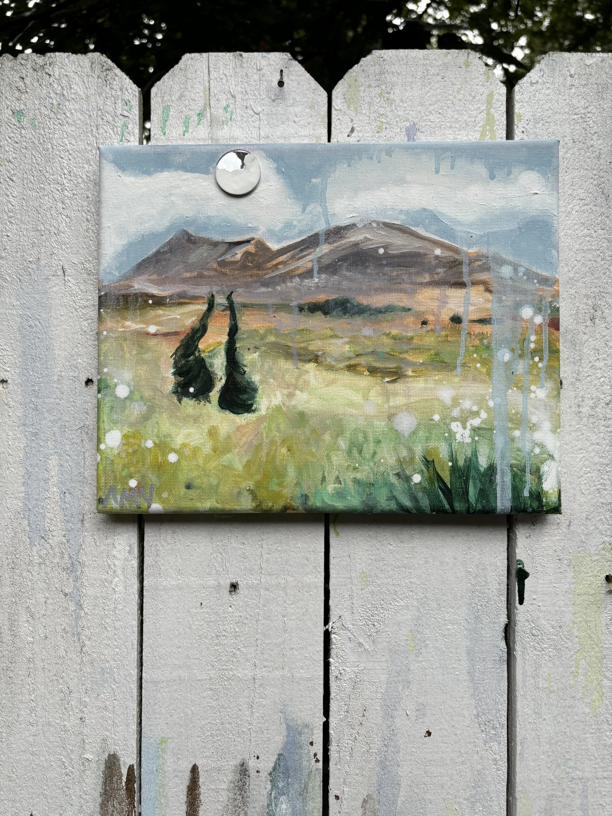 Watercolor landscape painting of mountains with cloudy sky, green field, trees, and two hanging objects in the foreground, mounted on a white wooden fence.