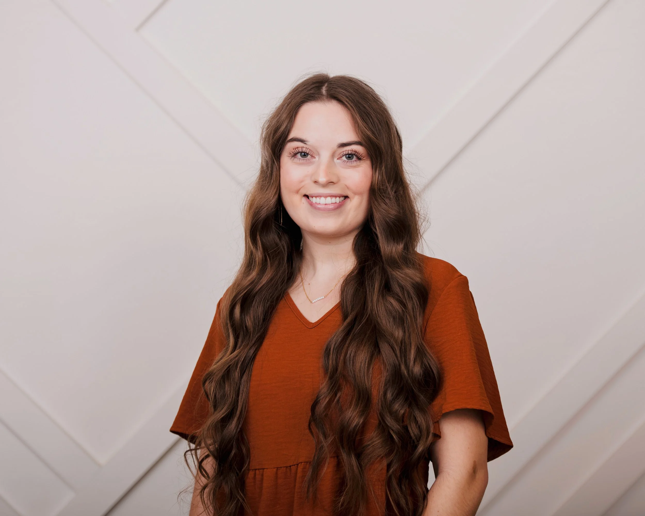 A young woman with long wavy brown hair, wearing a rust-colored shirt, standing indoors against a white wall with intersecting panel lines.