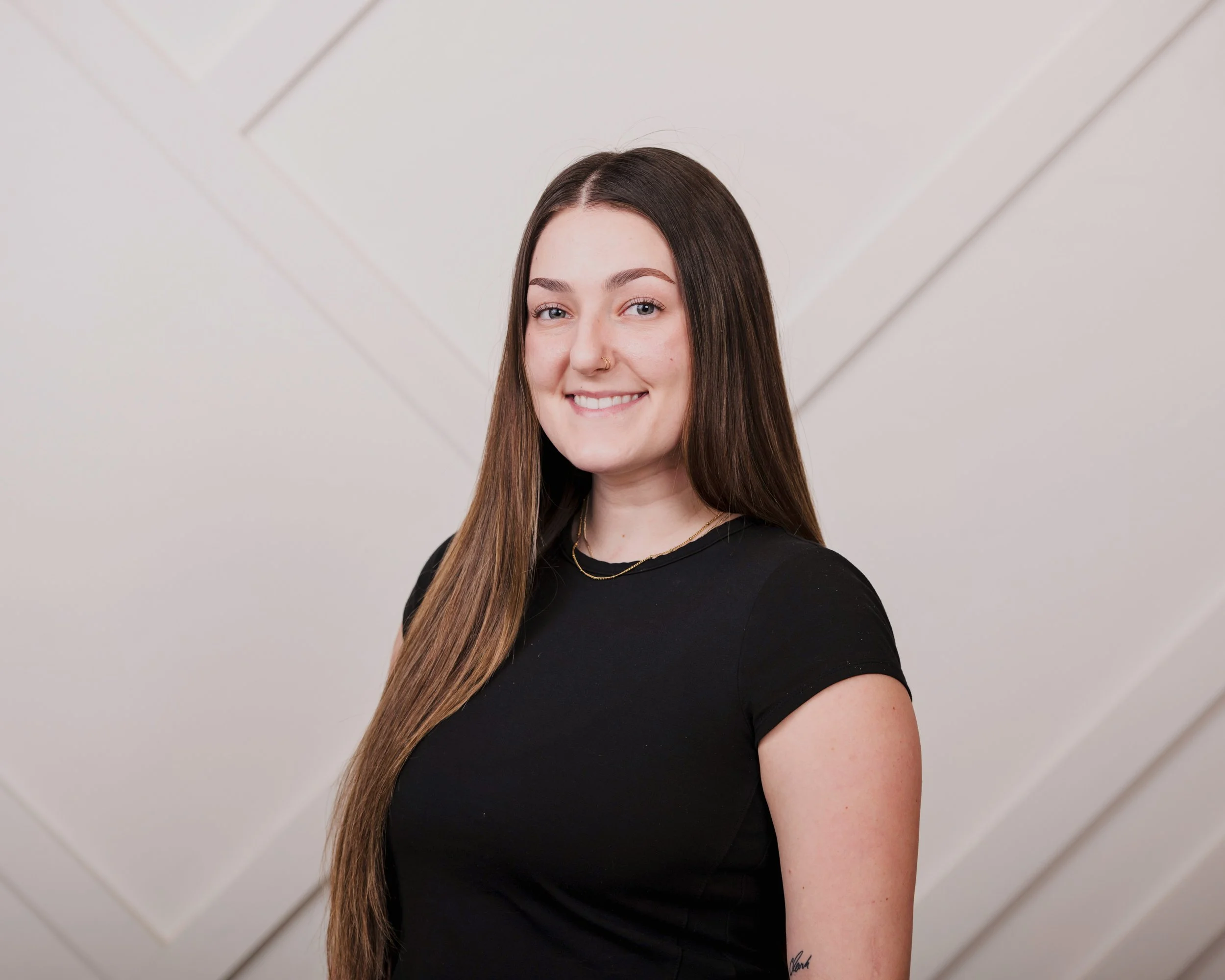 A portrait of a young woman with long brown hair, smiling, wearing a black top and a gold necklace, standing in front of a neutral background with geometric lines.