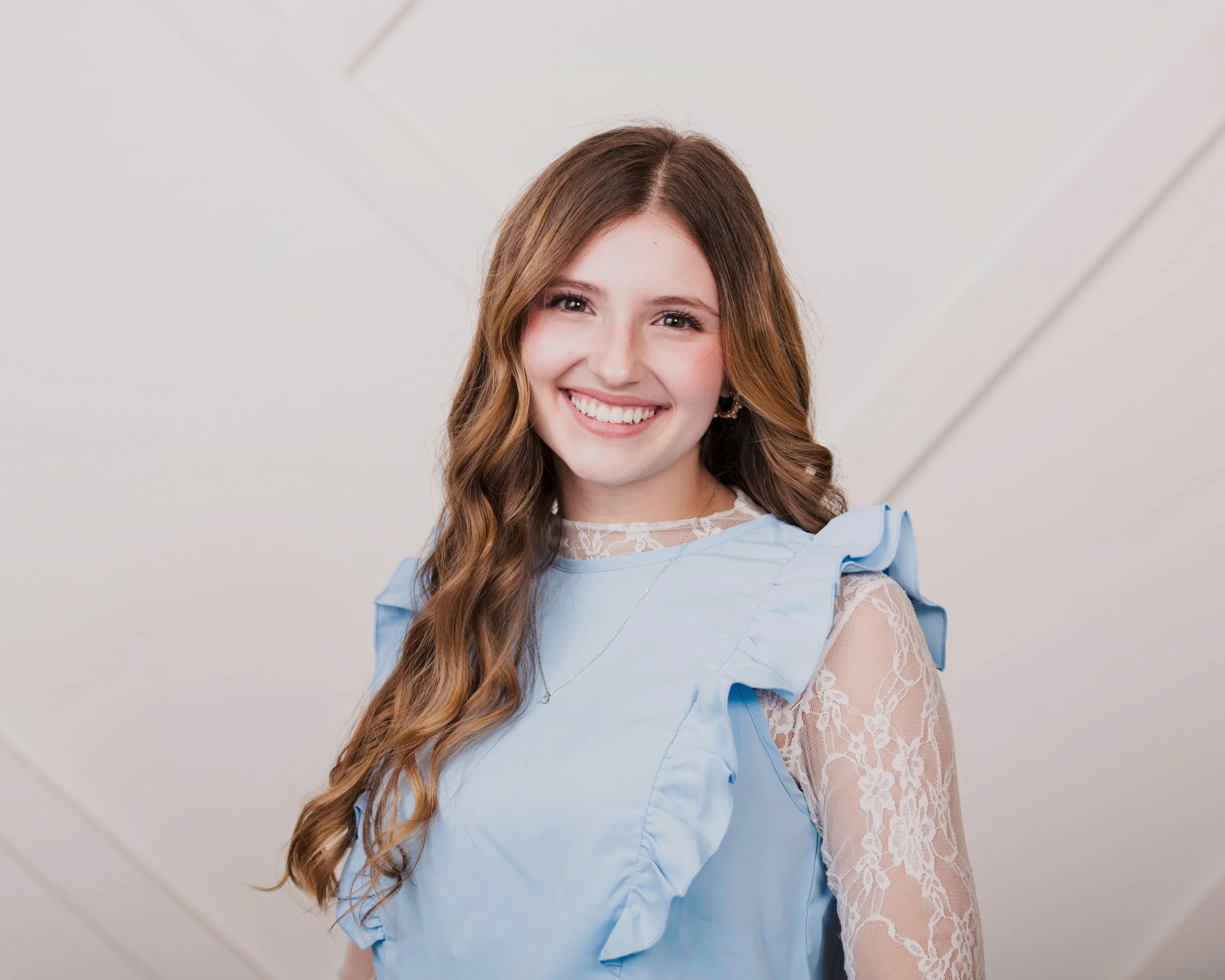 A young woman with long, wavy brown hair and a bright smile, wearing a light blue dress with lace details on the sleeves, standing against a plain white background.