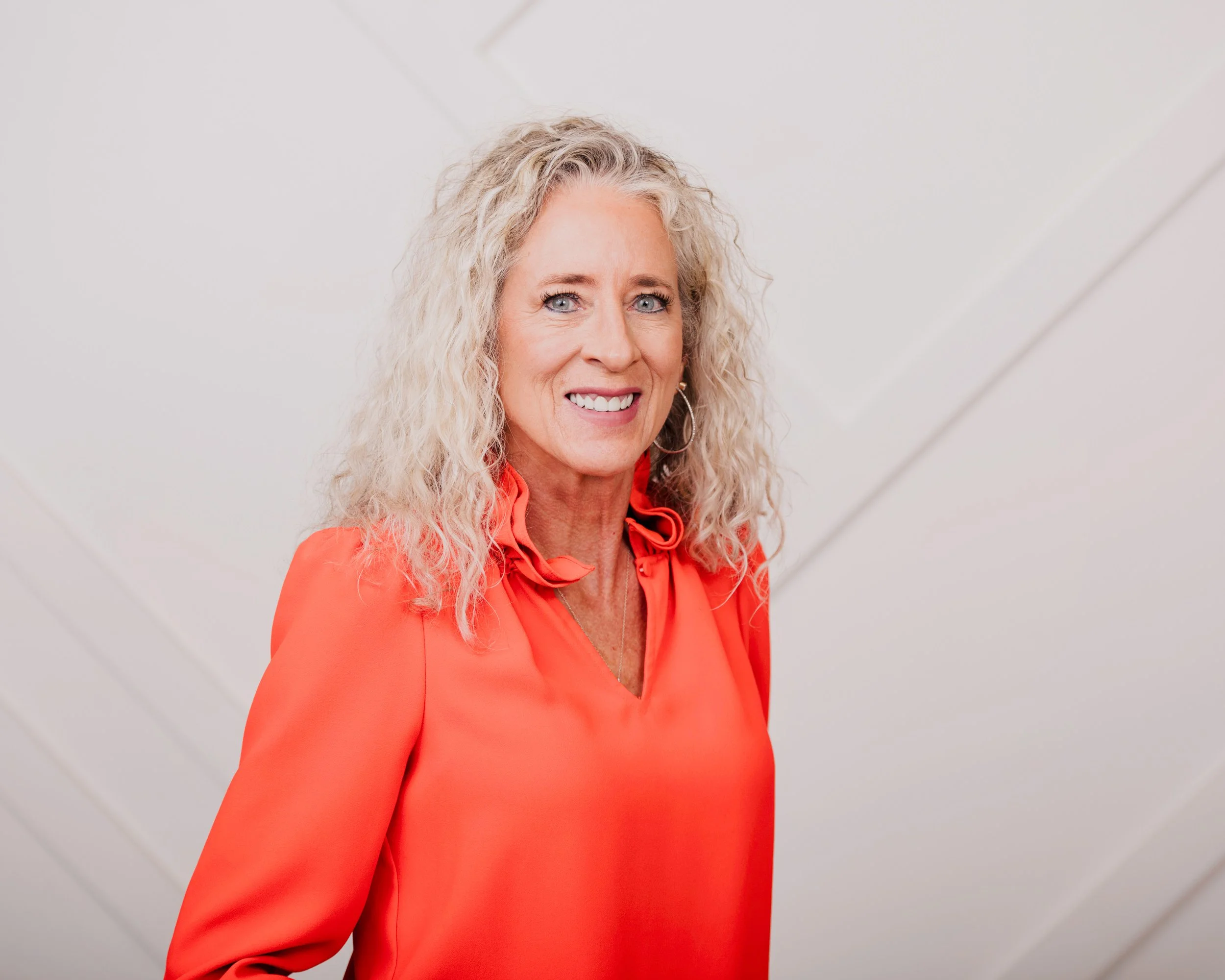 A woman with curly blonde hair and blue eyes smiling, wearing a bright orange blouse in front of a plain light gray background.