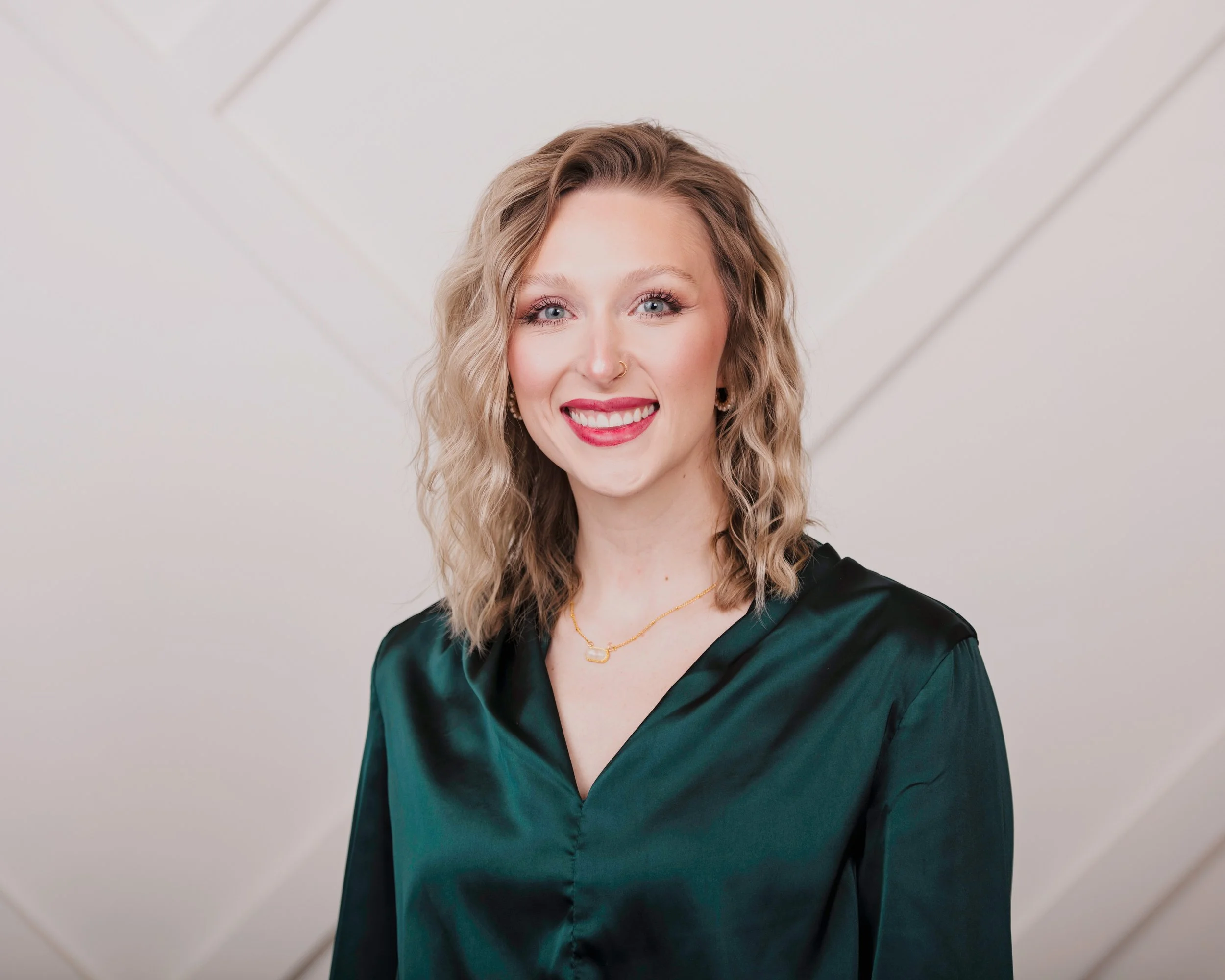 A woman with wavy blonde hair and blue eyes, smiling, wearing a dark green satin dress, a gold necklace, and a septum nose ring, standing against a light-colored background.