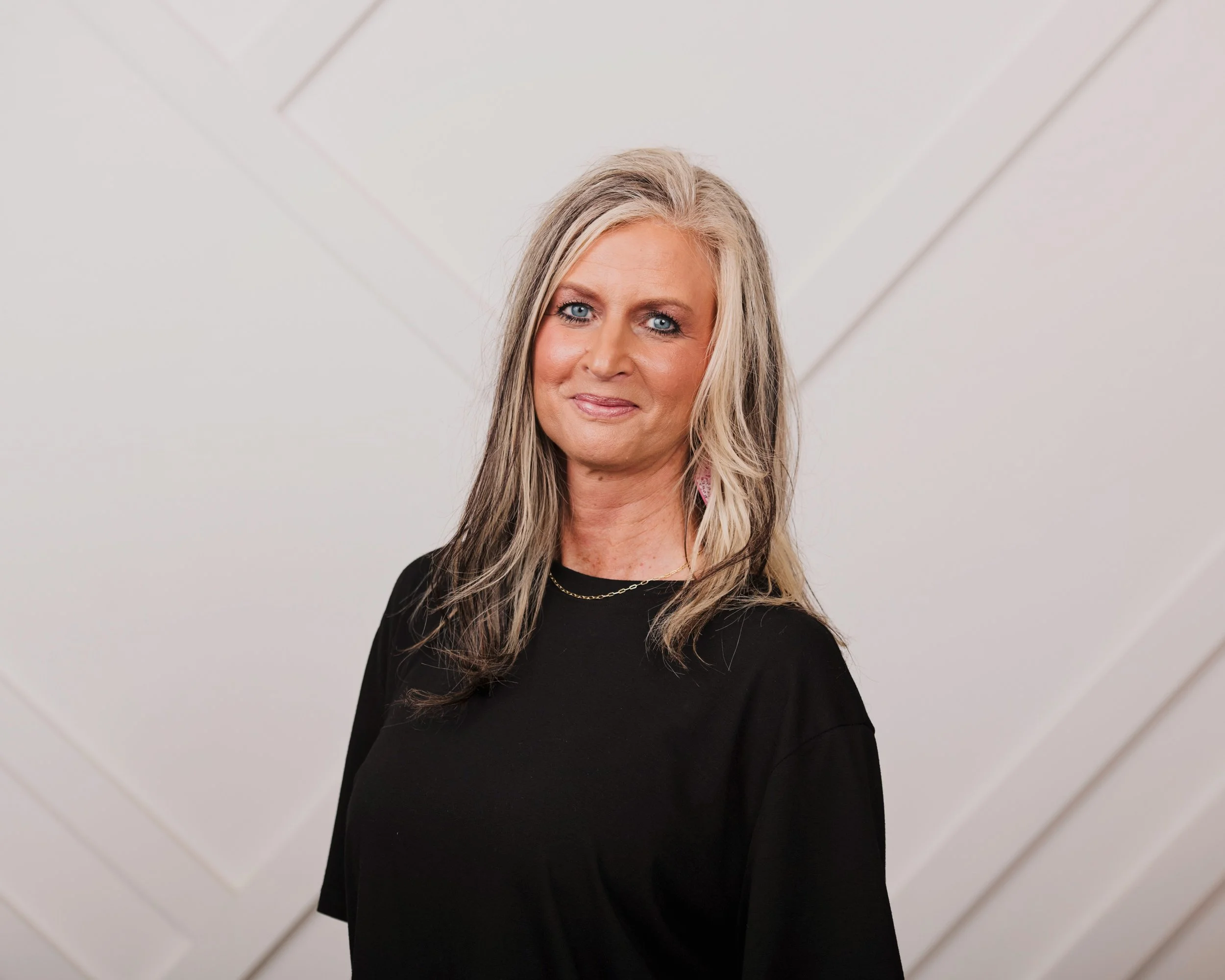 A middle-aged woman with long blonde hair and blue eyes wearing a black top, standing in front of a light-colored, tiled wall.