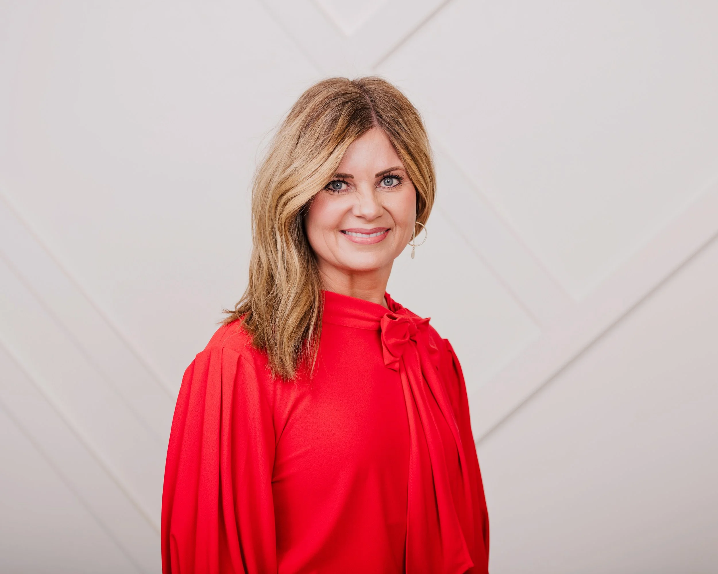 A woman with long, wavy blonde hair and blue eyes is smiling and wearing a bright red dress with a large bow detail on the shoulder. She is standing against a plain, white wall.