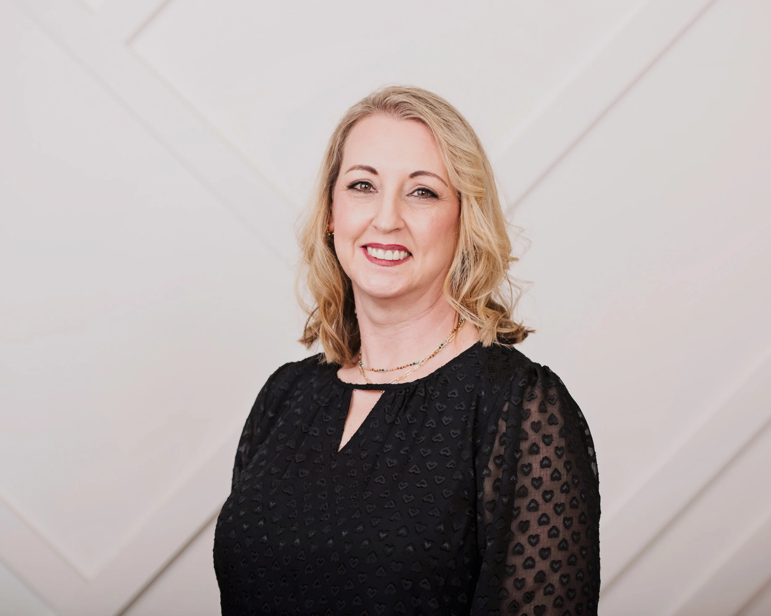 A woman with blonde wavy hair smiling, wearing a black dress with sheer sleeves decorated with a heart pattern, standing in front of a white wall.