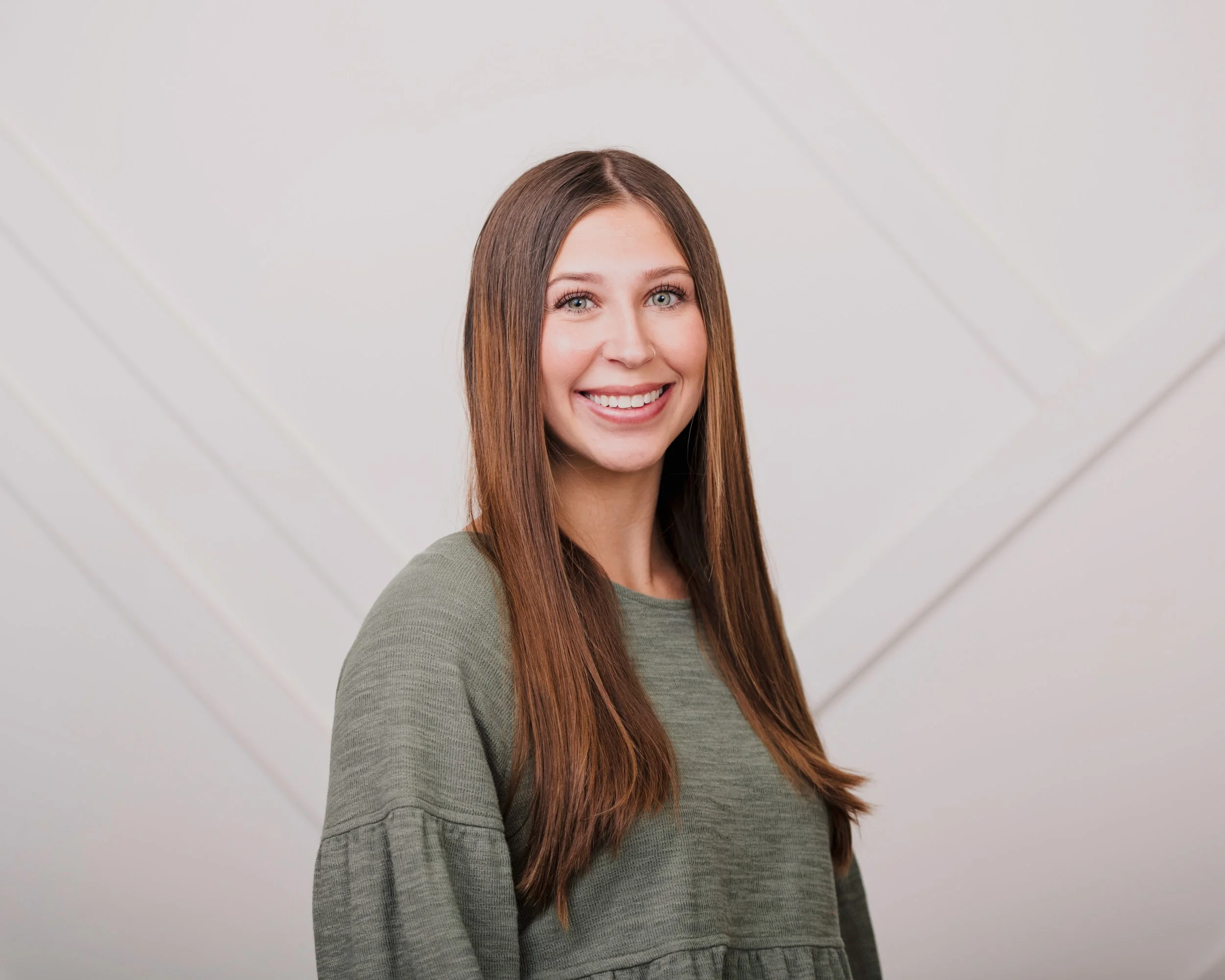 A young woman with long brown hair, blue eyes, and a nose piercing, smiling while looking at the camera, wearing a gray long-sleeve shirt, standing against a plain light-colored background.