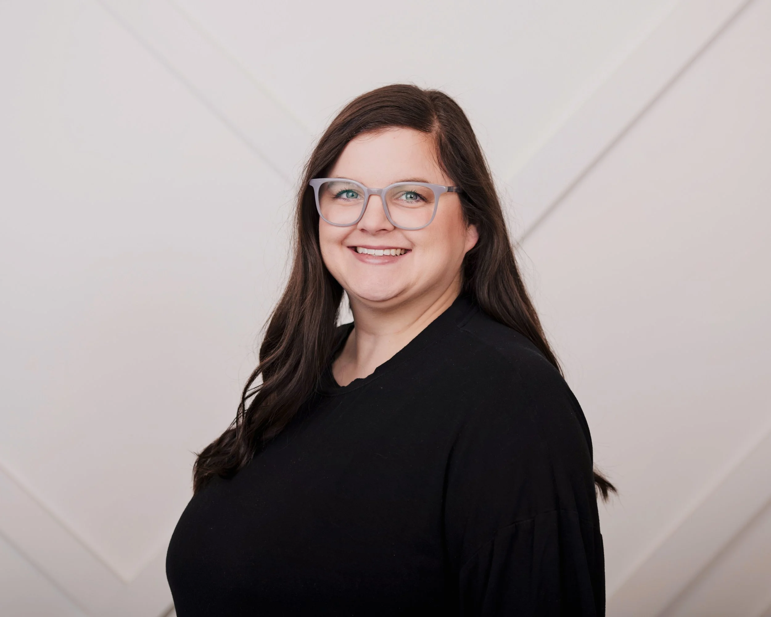 A woman with long dark hair, wearing glasses with clear frames, smiling, and dressed in a black top, against a plain light background.