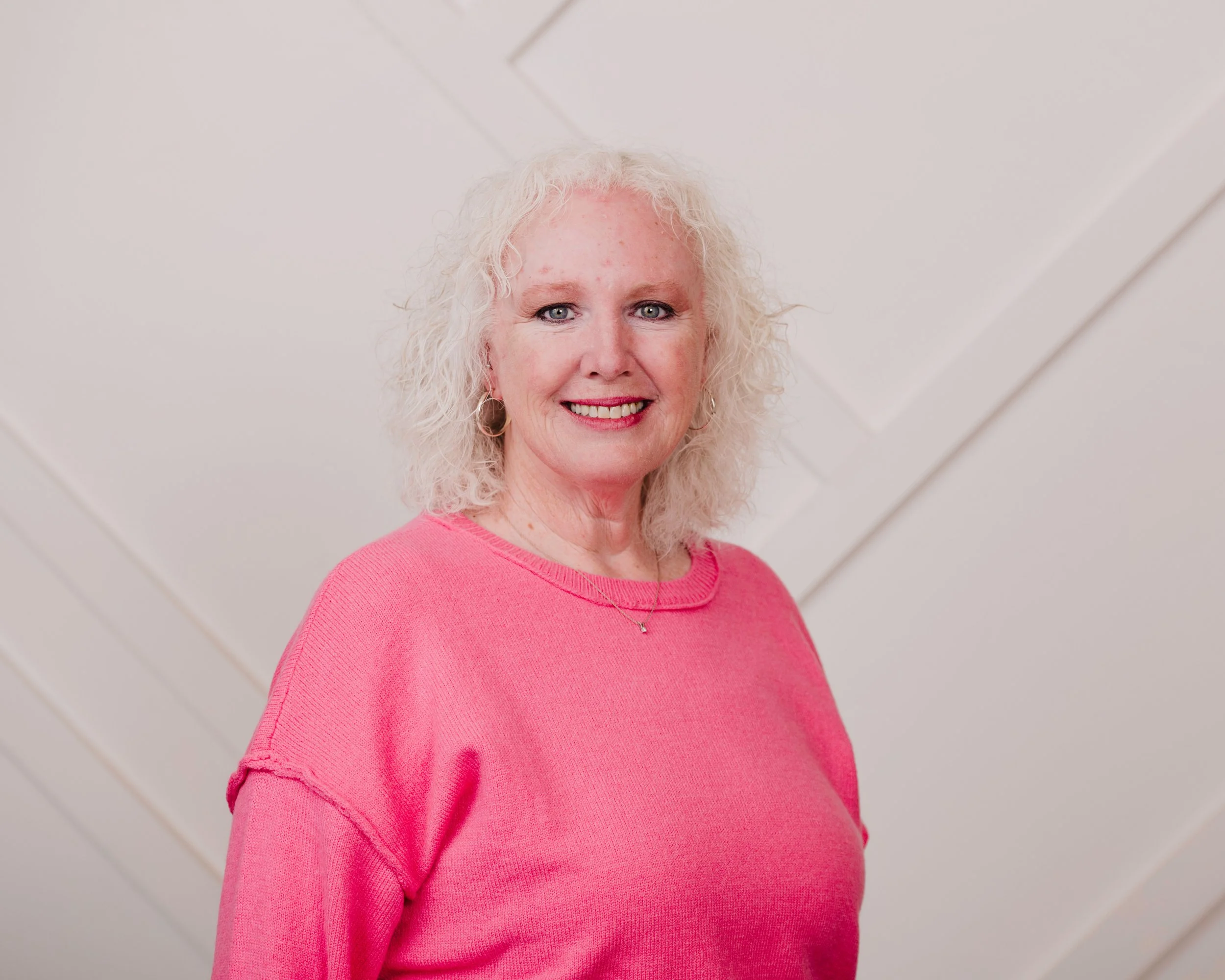 A middle-aged woman with curly white hair wearing a pink sweater, smiling at the camera, standing against a plain white wall.