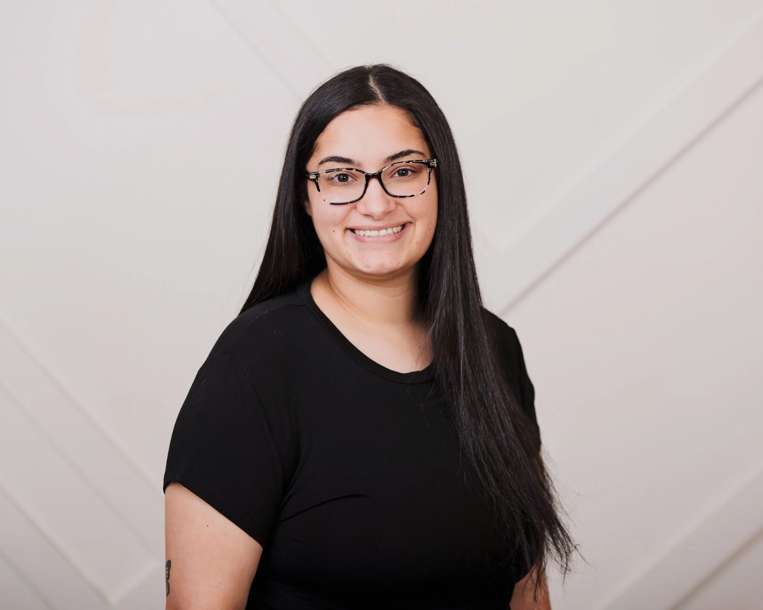 A woman with long dark hair wearing glasses and a black shirt, smiling in front of a plain white wall.