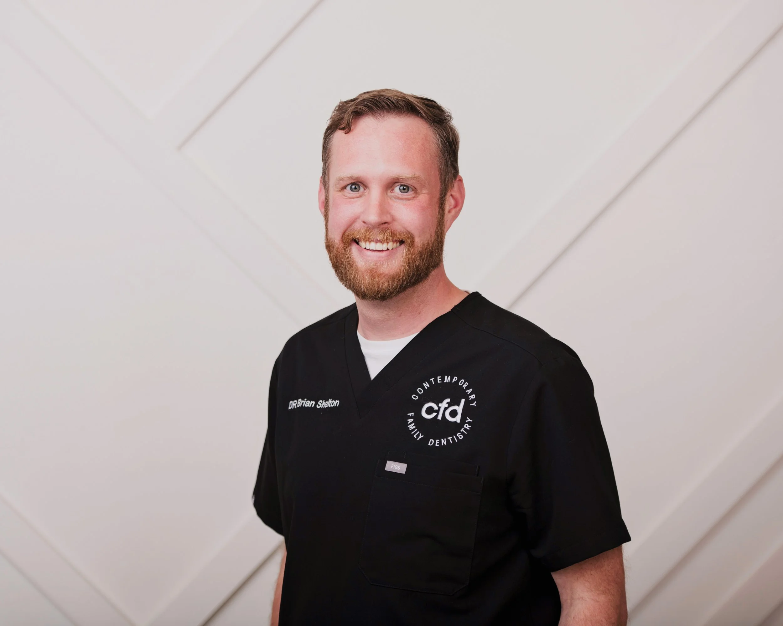 A smiling man with a beard and mustache, wearing a black dental scrubs top, standing in front of a neutral background.