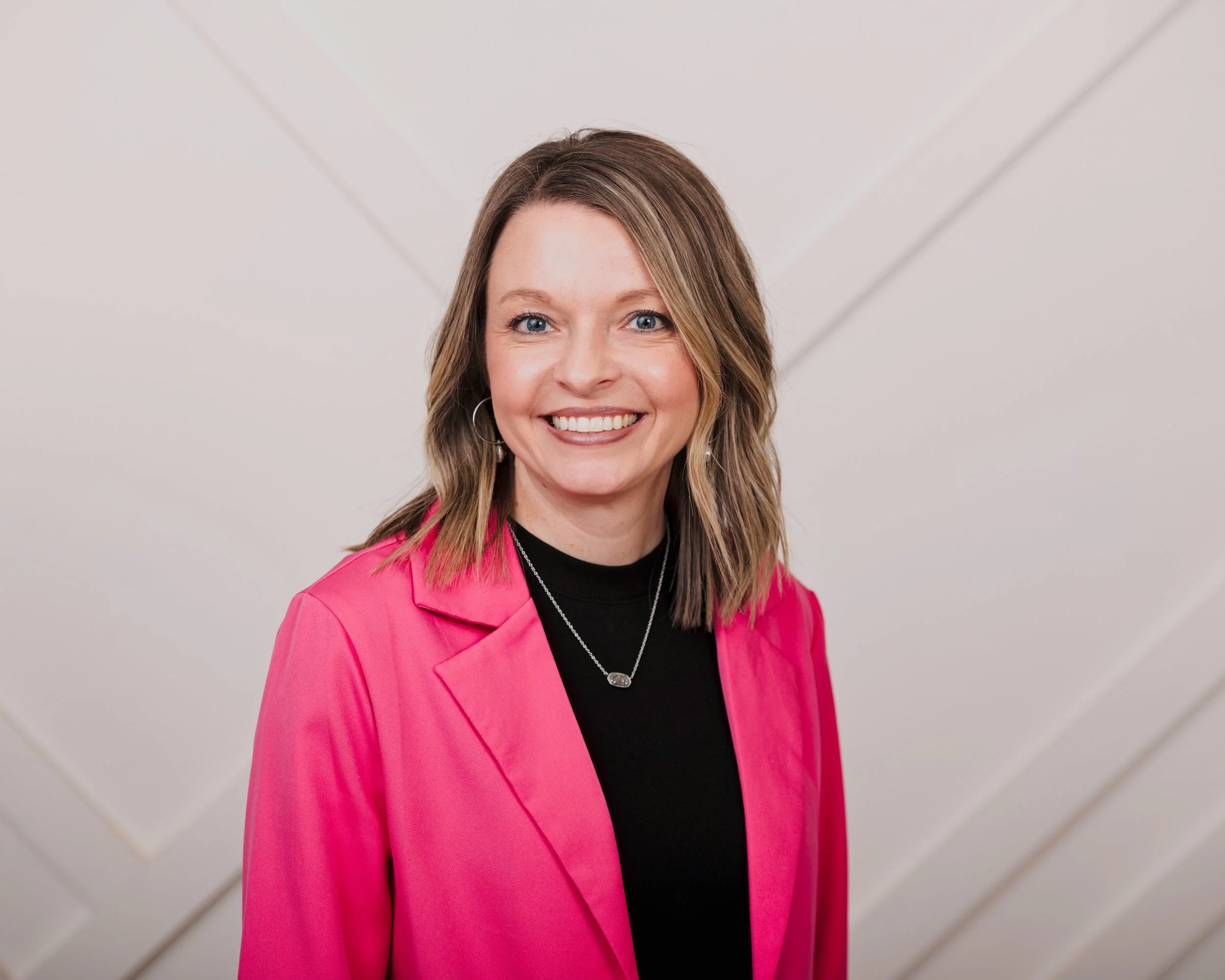 A woman with shoulder-length wavy hair, wearing a bright pink blazer over a black top, smiling at the camera against a neutral background with faint diagonal lines.