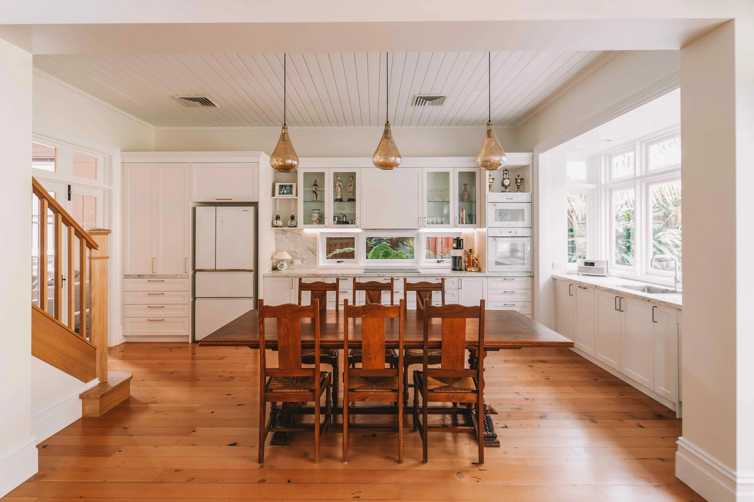 A kitchen with white cabinets, a wooden dining table with six chairs, wooden flooring, three hanging pendant lights, and large windows allowing natural light.