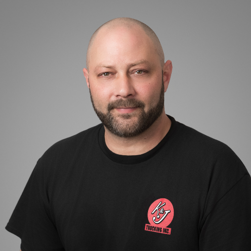 A middle-aged man with short, light-colored hair and blue eyes, wearing a gray work shirt with two name patches that read 'Keith' and initials 'K&J' on a gray background. He is looking at the camera with a slight smile against a gray background.