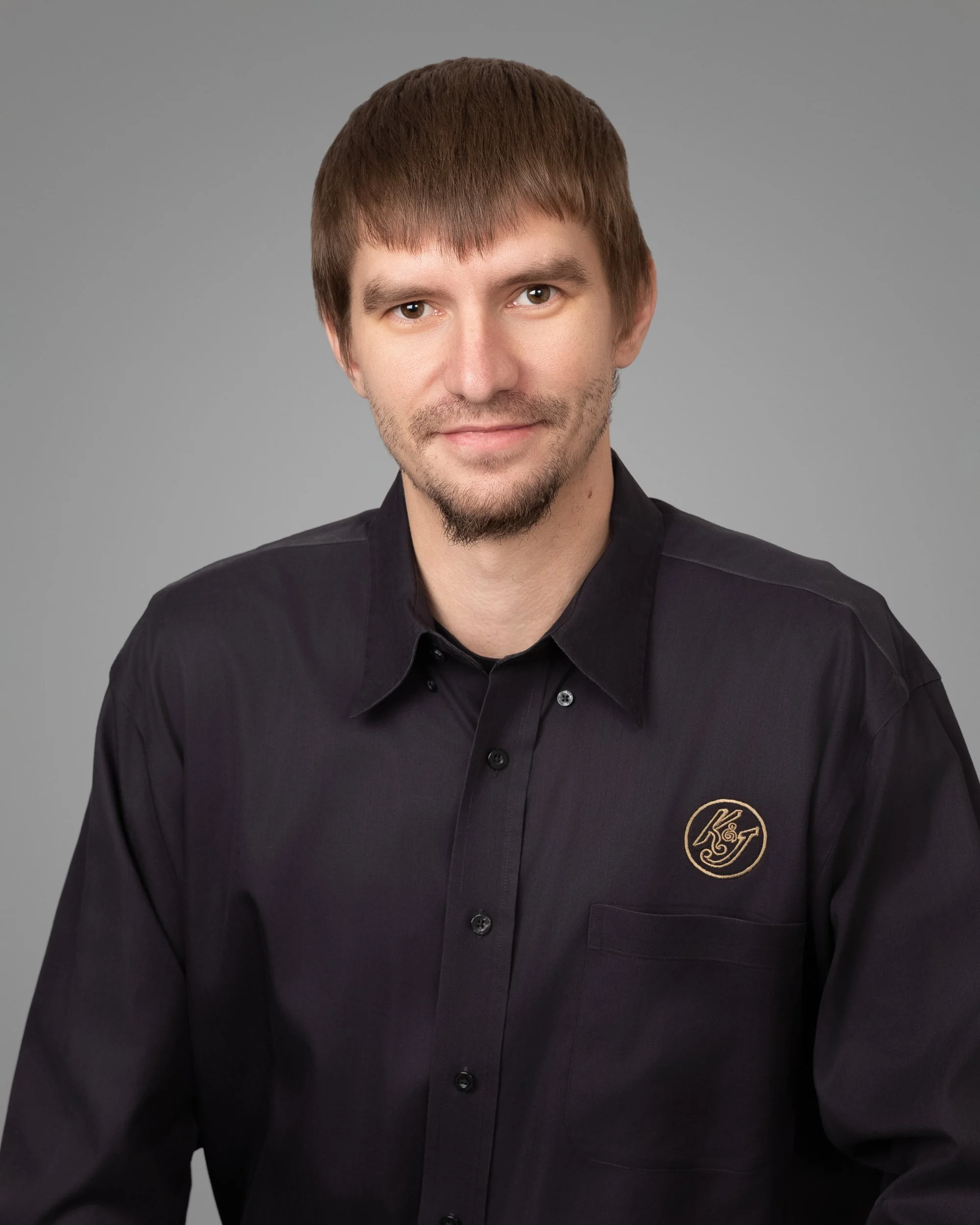 A man with brown hair, a beard, and a smile wearing a black button-up shirt with a gold embroidered logo on the chest, against a gray background.