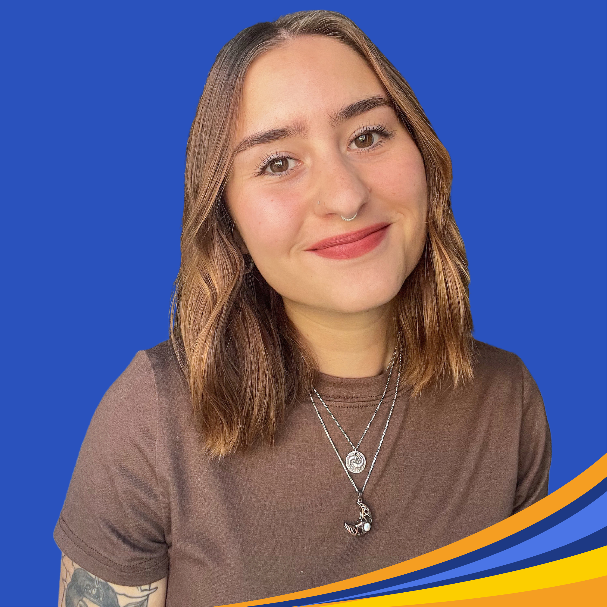 smiling woman with brown wavy hair in brown t-shirt with nose piercing and tattoo on arm