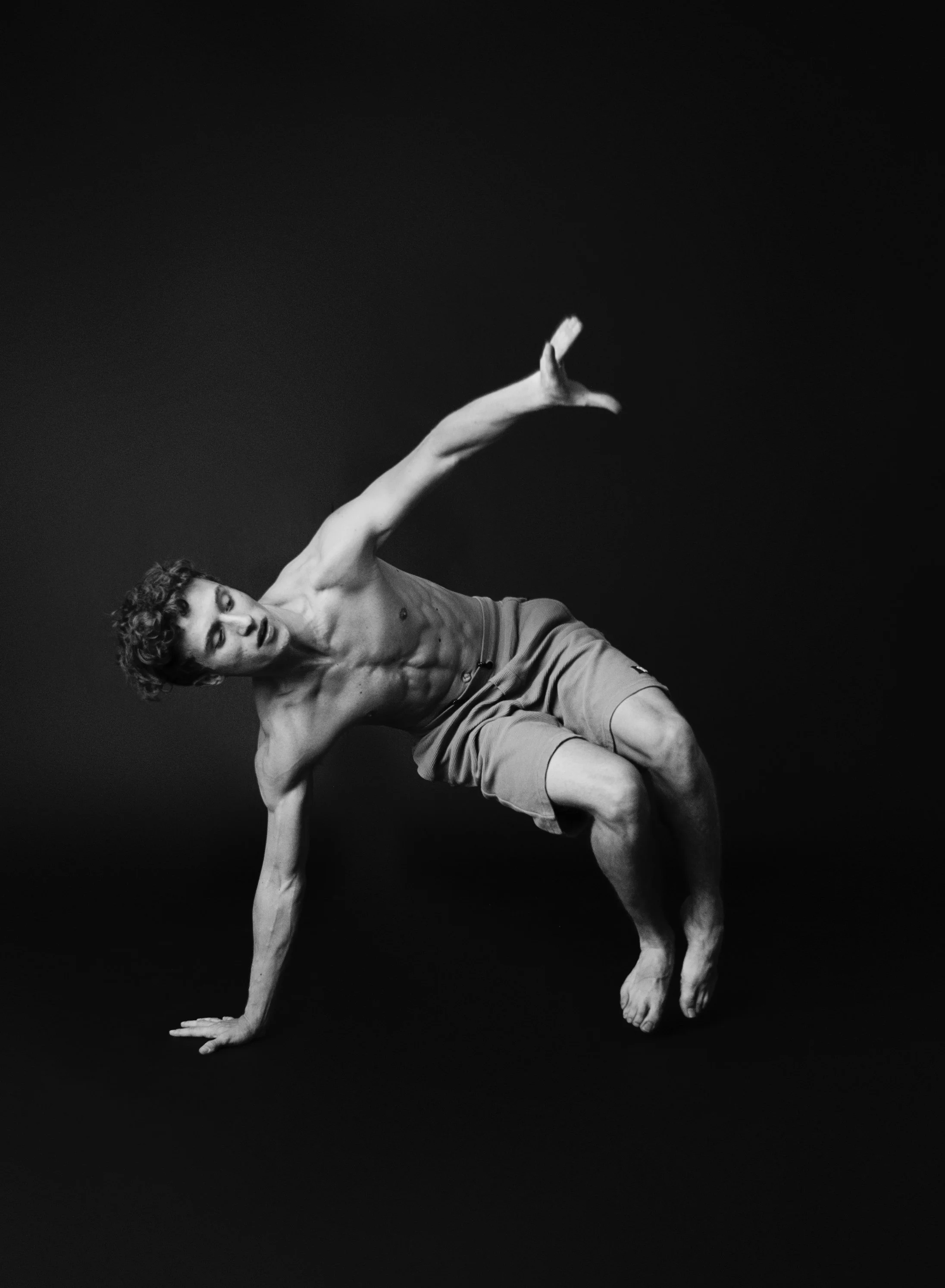 A shirtless young man with curly hair performing a breakdance move against a dark background, balancing on one hand with legs bent and one arm extended outward.