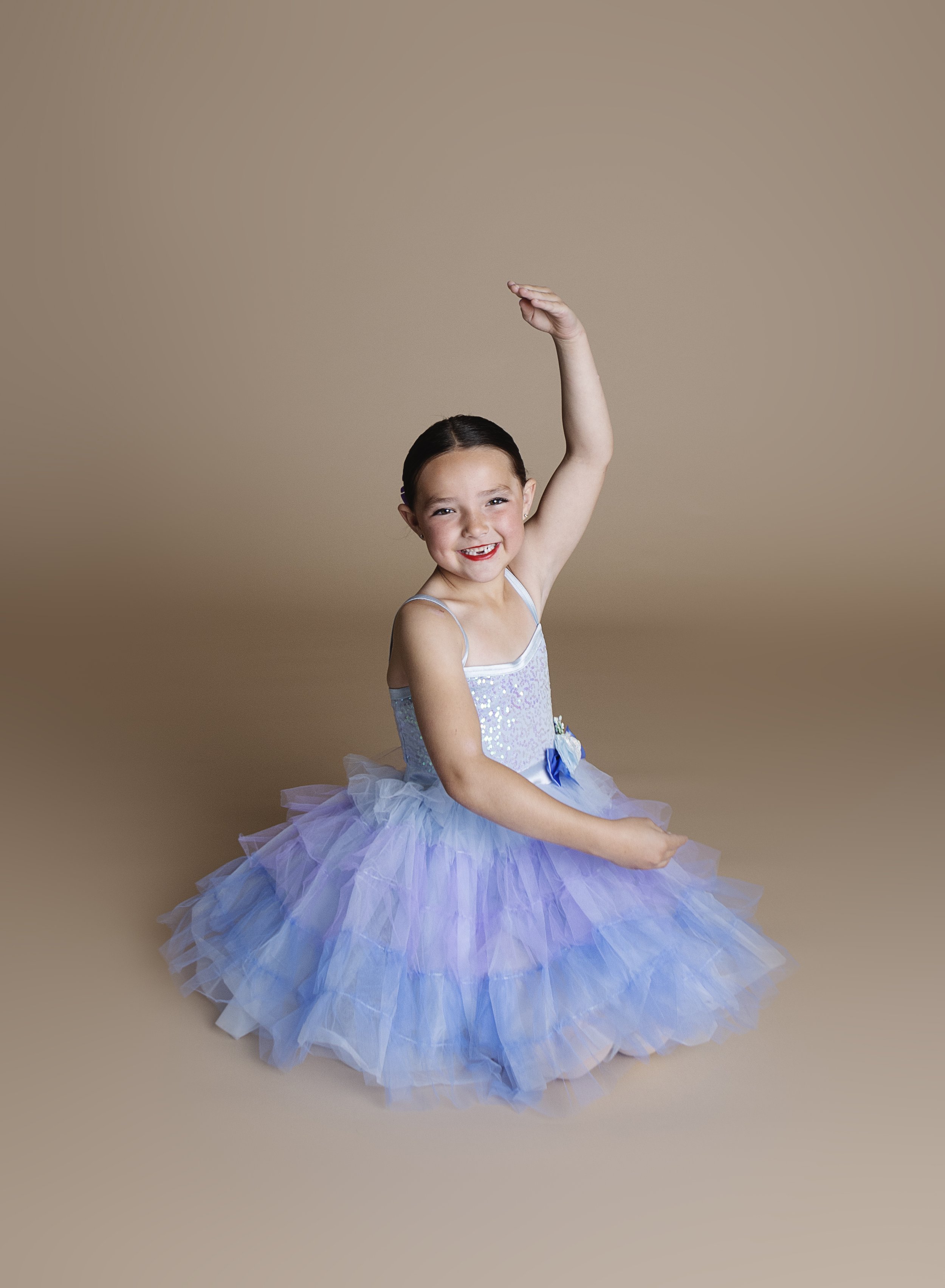 A young girl in a pastel blue and purple tutu dress, smiling and sitting on the floor with one arm raised high.