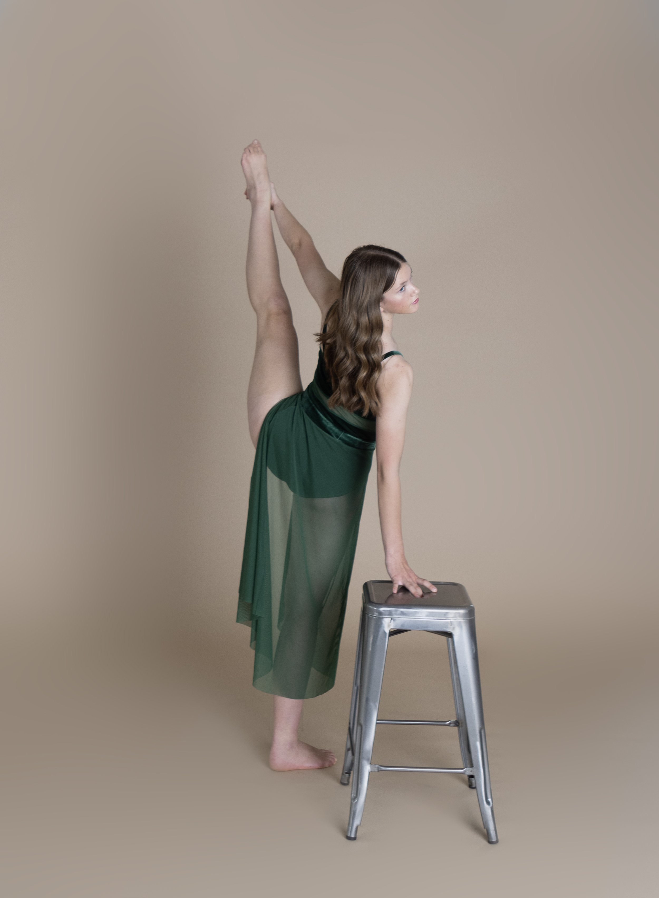 A woman in a green dress performing a standing split with her left foot on the high stool and her right hand on the stool for support. She is looking to her right with long wavy brown hair in a beige studio setting.