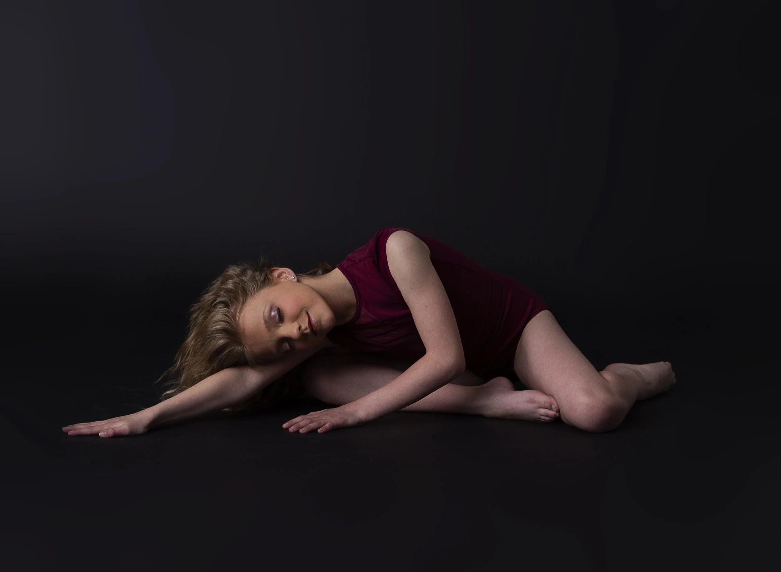 A woman with long hair, wearing a sleeveless dark red dress, is lying on her side on a black background, with her eyes closed and one arm stretched out on the floor.