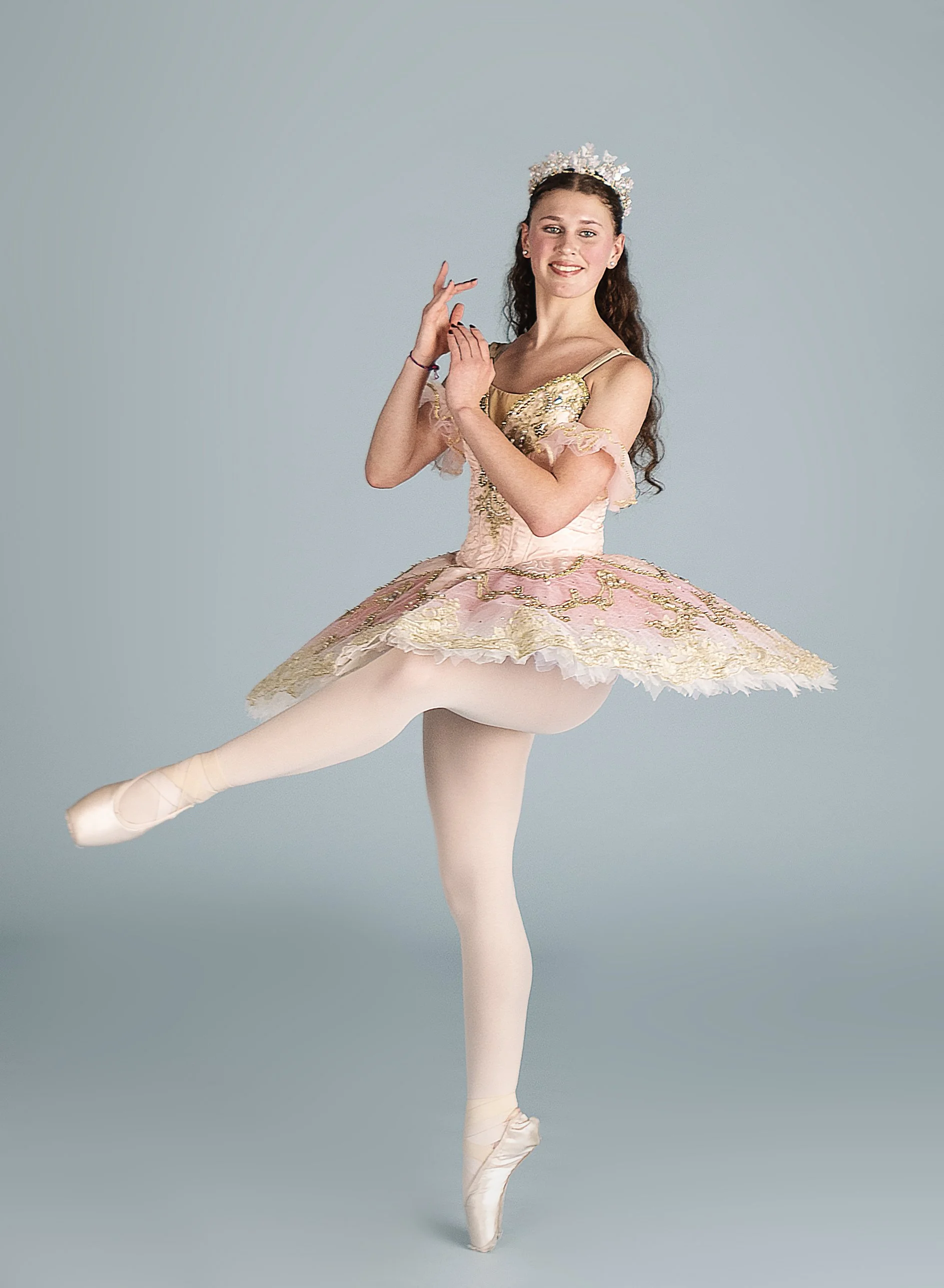 Ballet dancer in pink and gold tutu performing en pointe with one leg lifted, wearing a tiara, against a gray background.