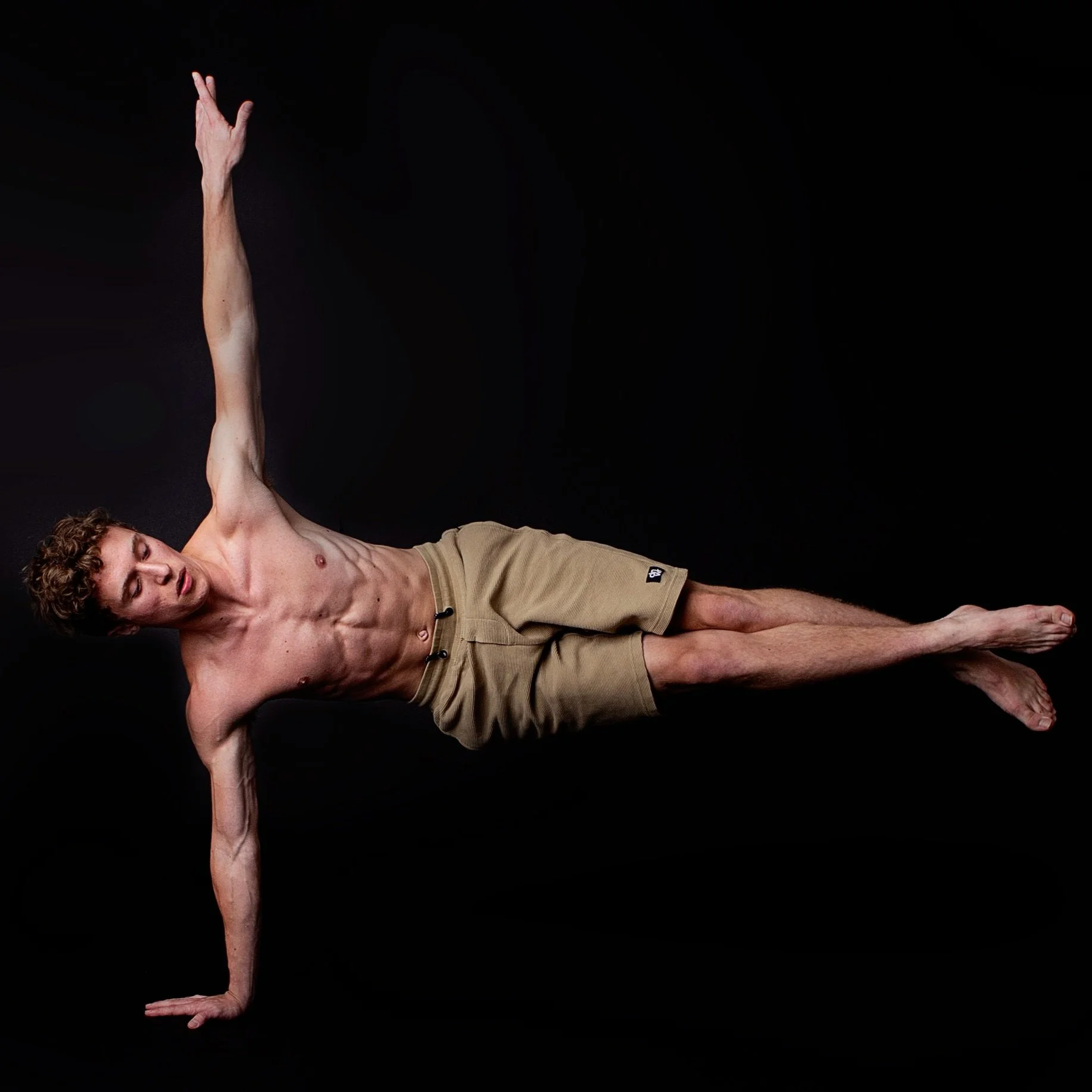 A young shirtless man with curly hair performing a side plank exercise against a black background, with his right hand on the ground and his left arm extended upward.