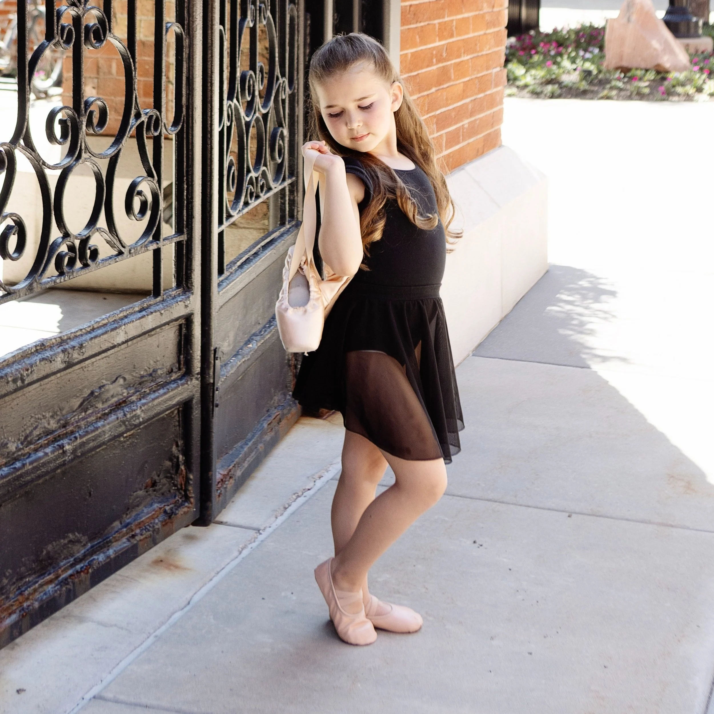 A young girl in a black dress and ballet shoes leaning against a black iron gate outside a brick building, holding a pale pink bag over her shoulder.