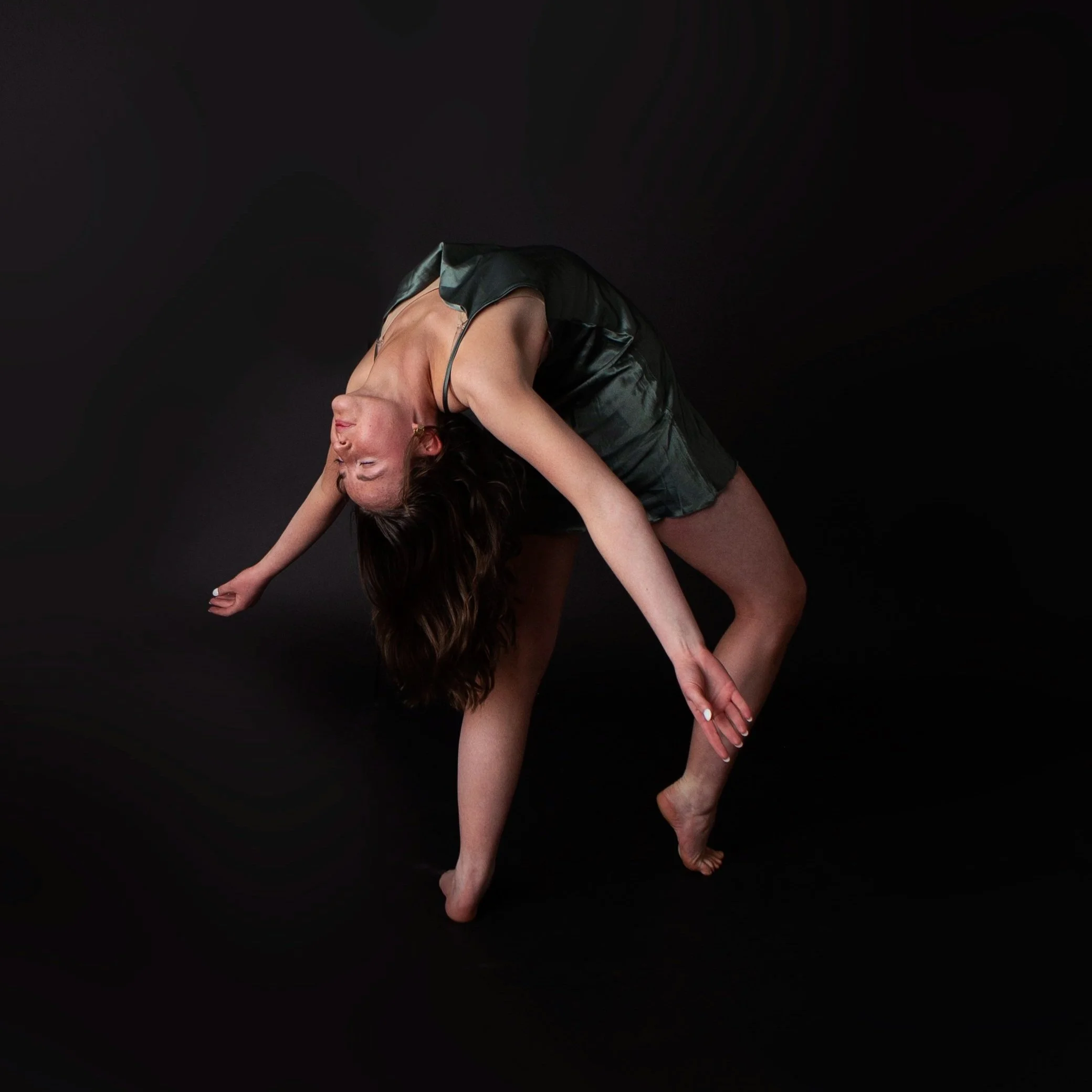 A woman in a black satin dress performing a yoga pose on a black background, bending forward with one hand touching the ground and the other arm extended diagonally.