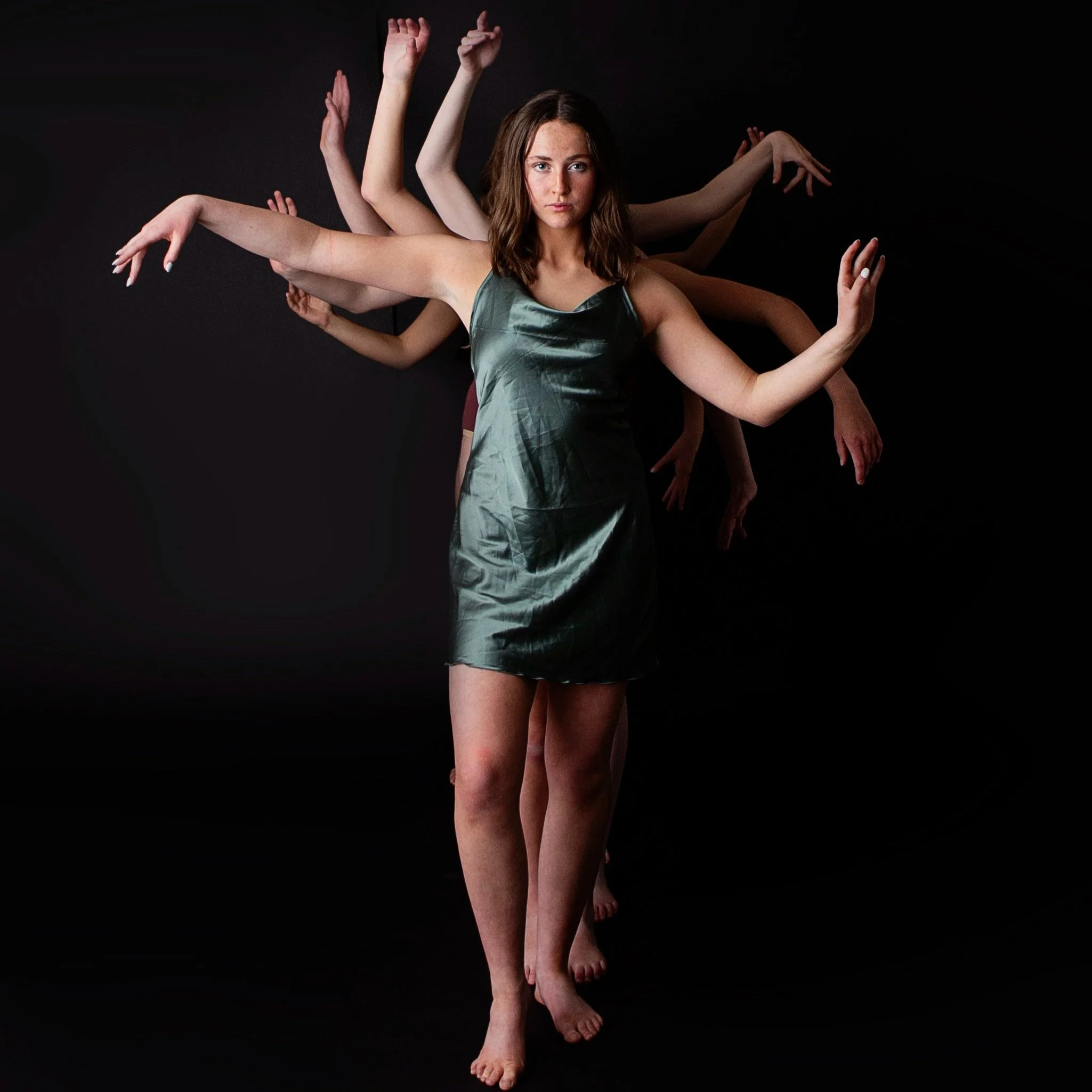 A woman in a green dress standing against a black background with multiple arms positioned around her, creating a visually surreal effect.