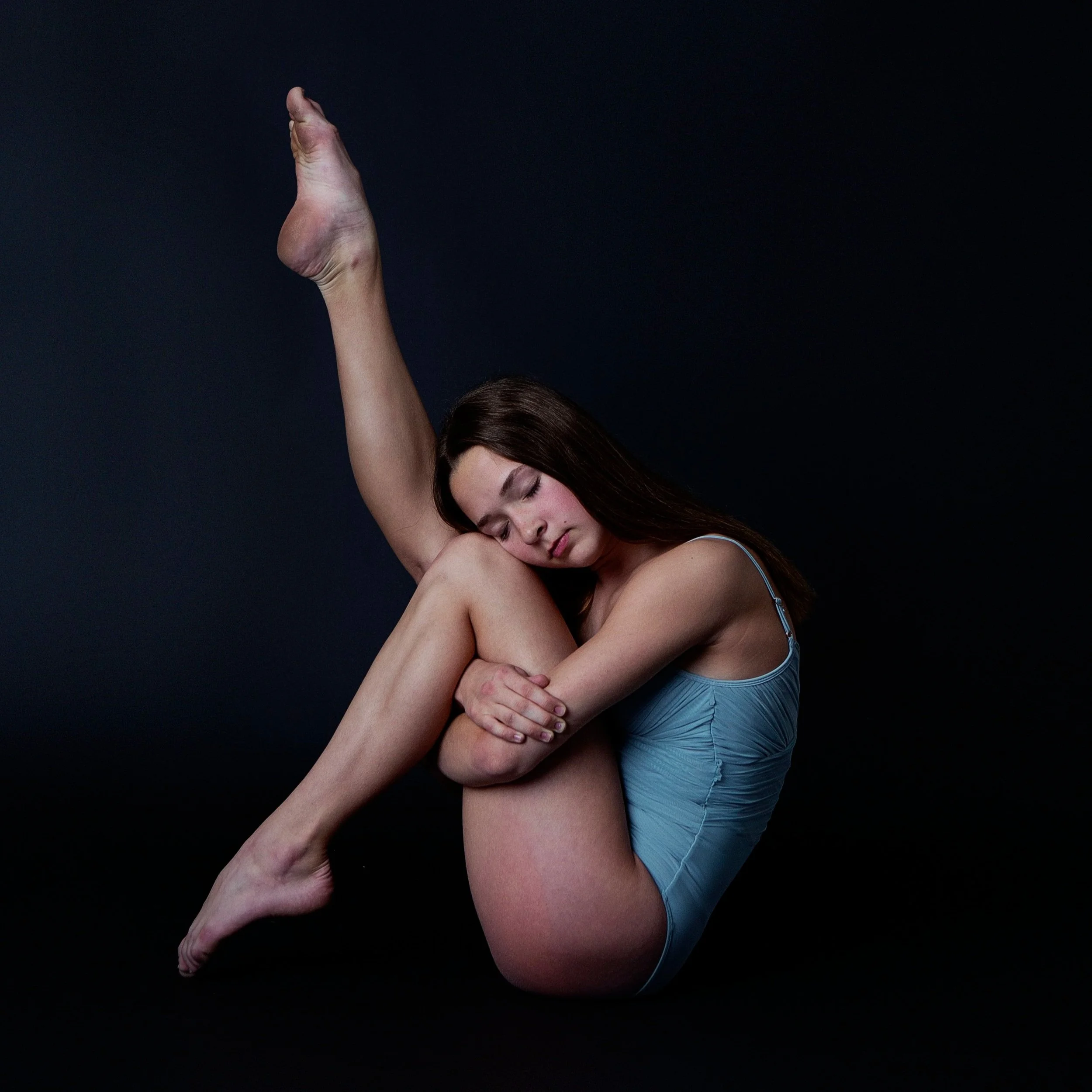 Young woman with long brown hair in a light blue tank top and shorts poses with her legs crossed and arms around her knees in a yoga pose, against a dark background.