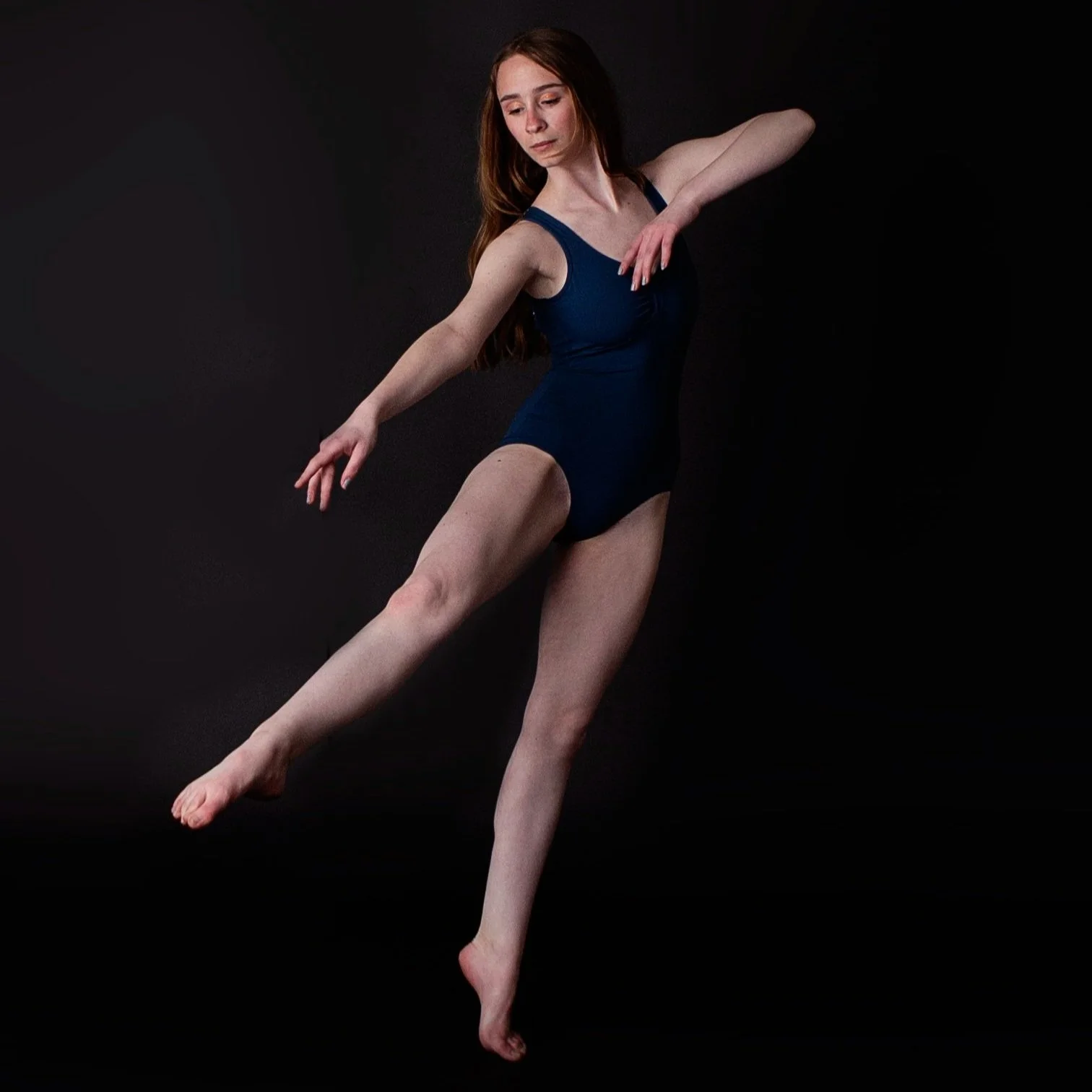 A young woman with long brown hair, wearing a blue leotard, is performing a dance pose against a black background, with her arms and legs extended gracefully.